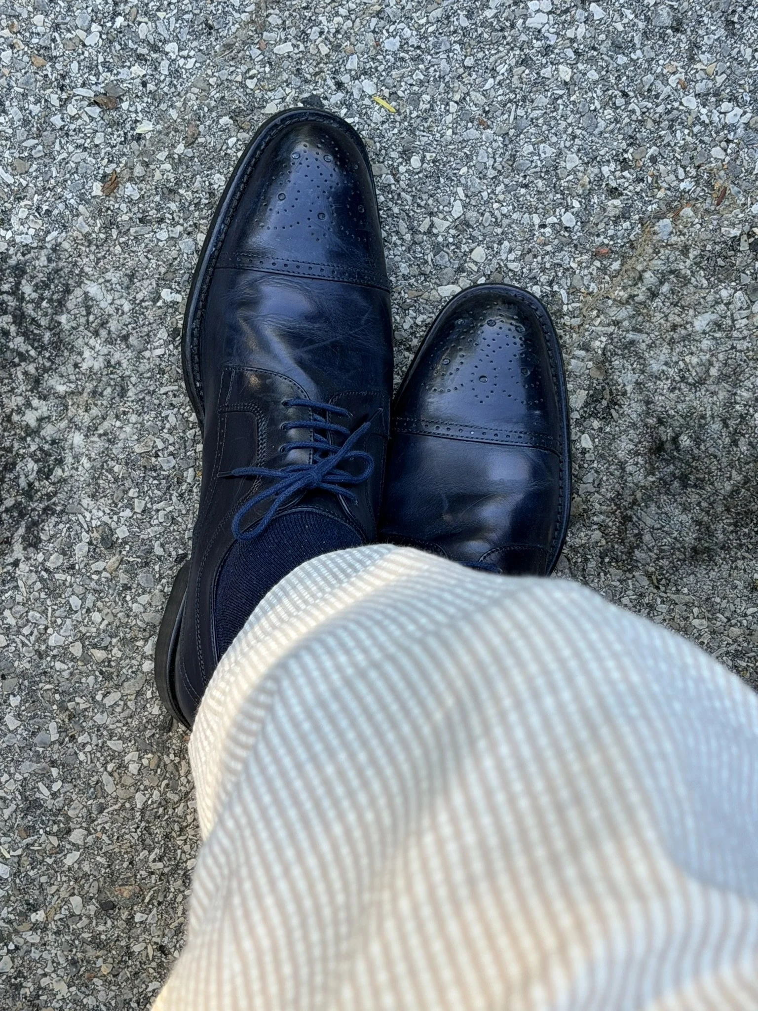 A person's feet in black leather dress shoes standing on a gravel surface, wearing beige striped pants.