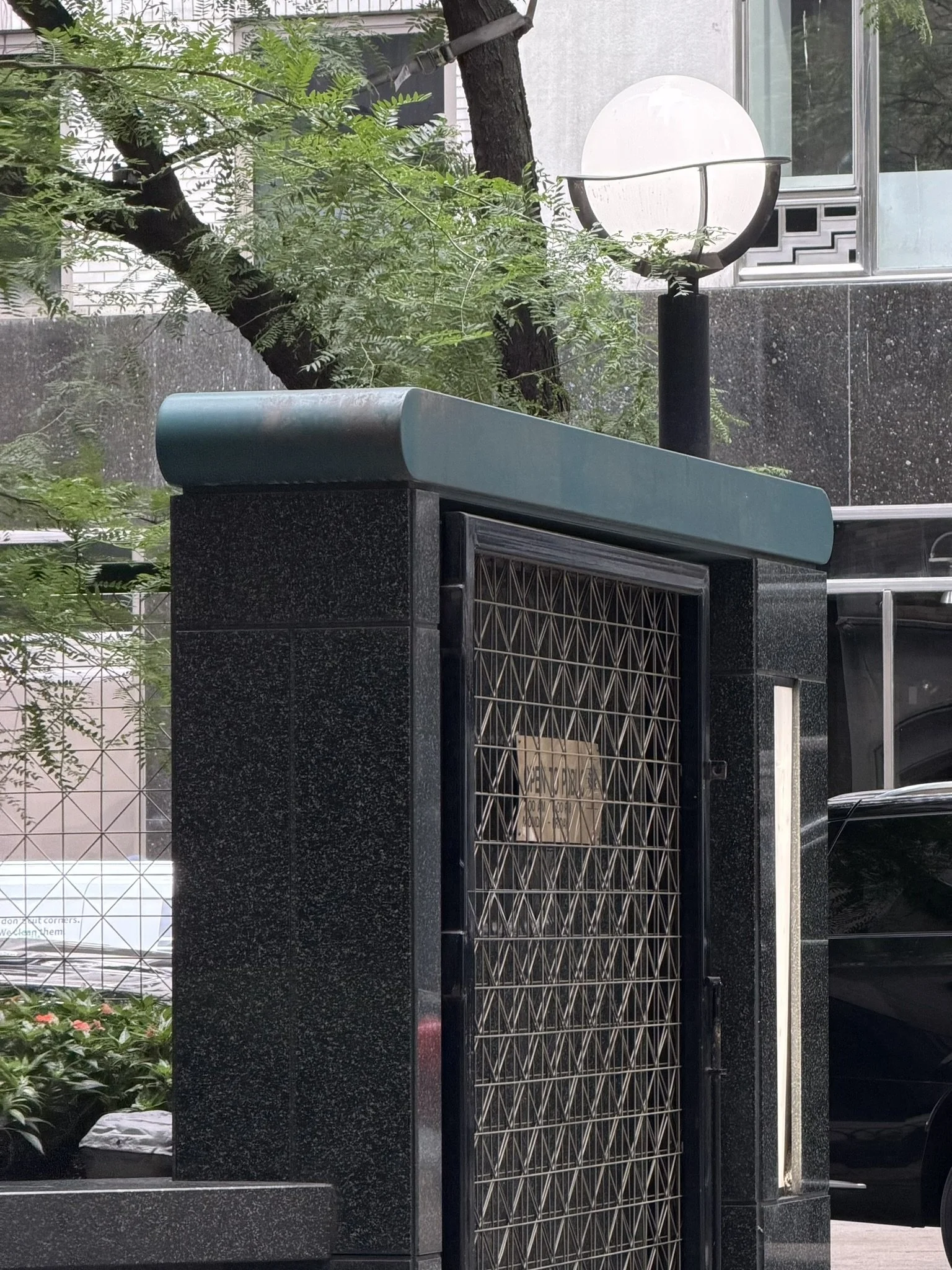 A black granite entrance with a metal gate and an overhead lampshade, with greenery including trees and plants nearby.