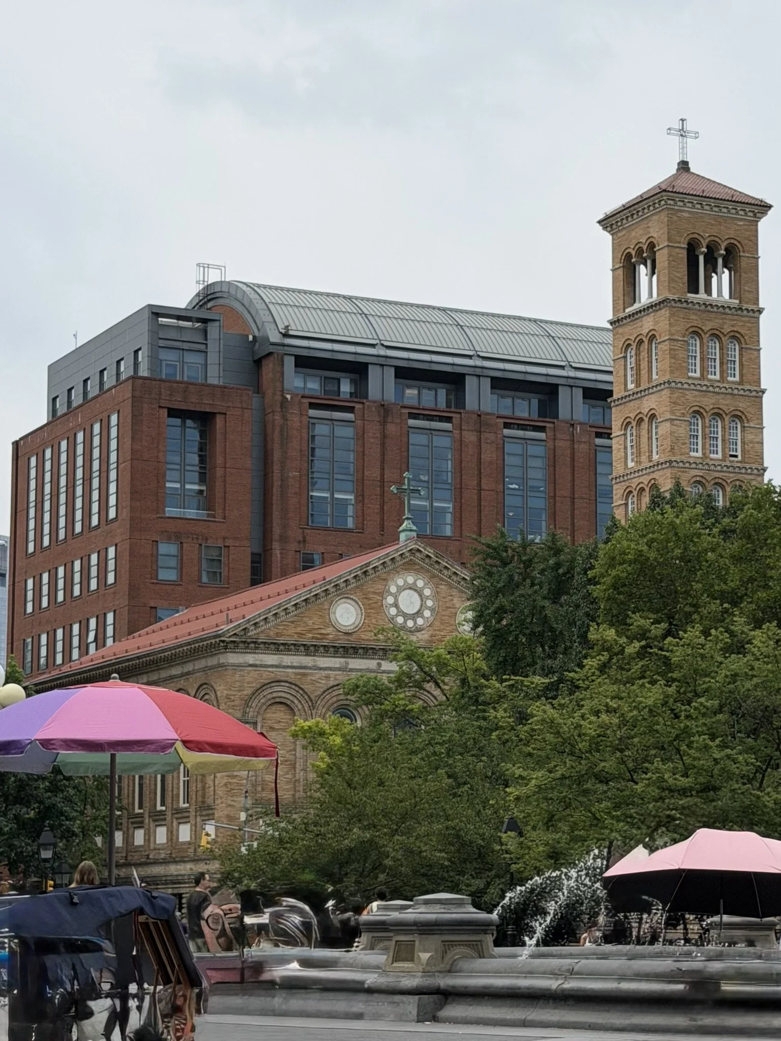 View of a city park with umbrellas, trees, and a fountain in the foreground, and historic and modern buildings, including a church with a clock and a brick bell tower, in the background.
