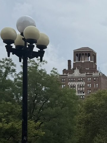 A vintage-style street lamp with five globes, with a city building and trees in the background under a cloudy sky.