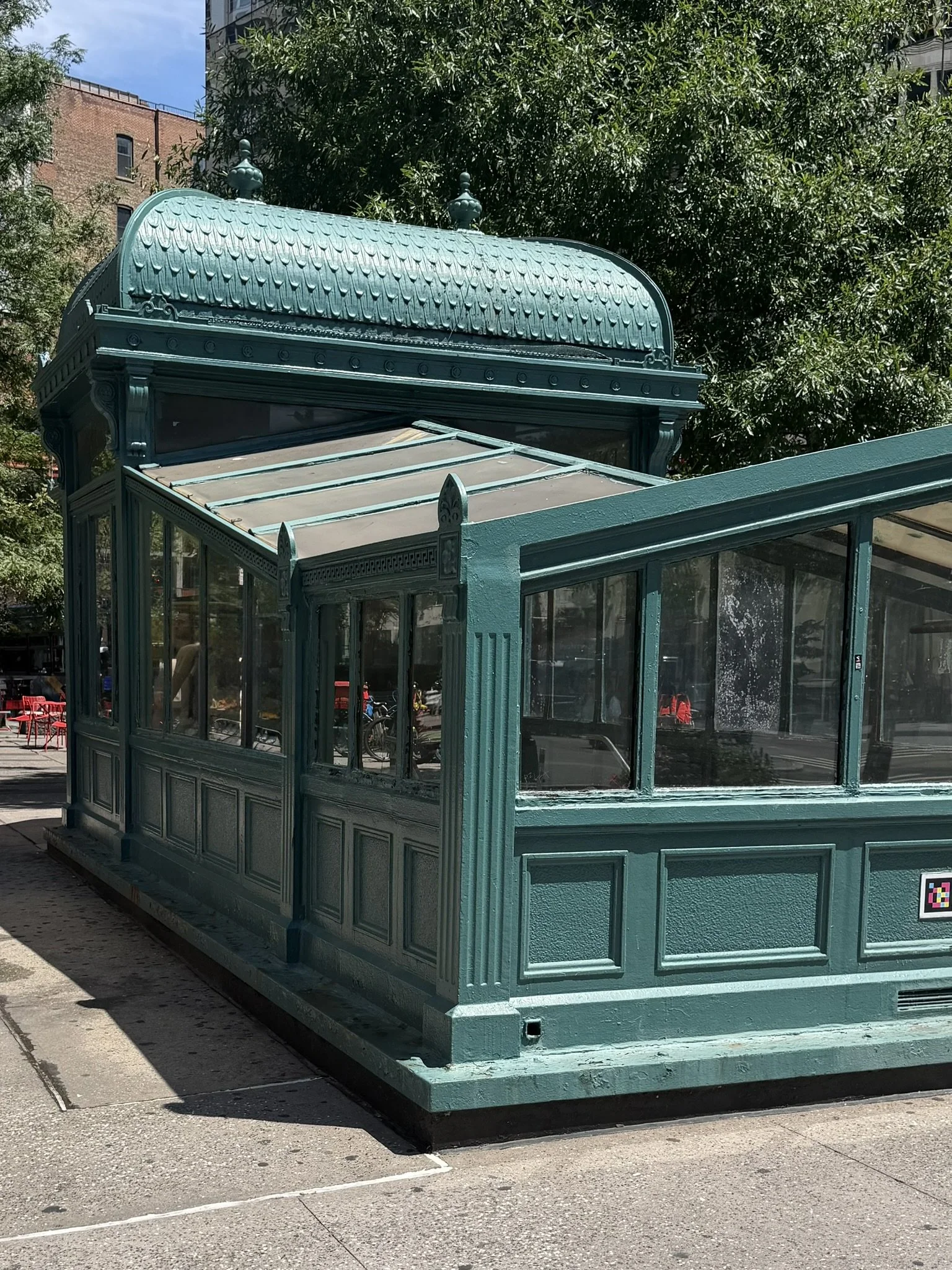 A vintage teal-colored glass and metal greenhouse or kiosk on a city sidewalk with trees and brick buildings in the background.