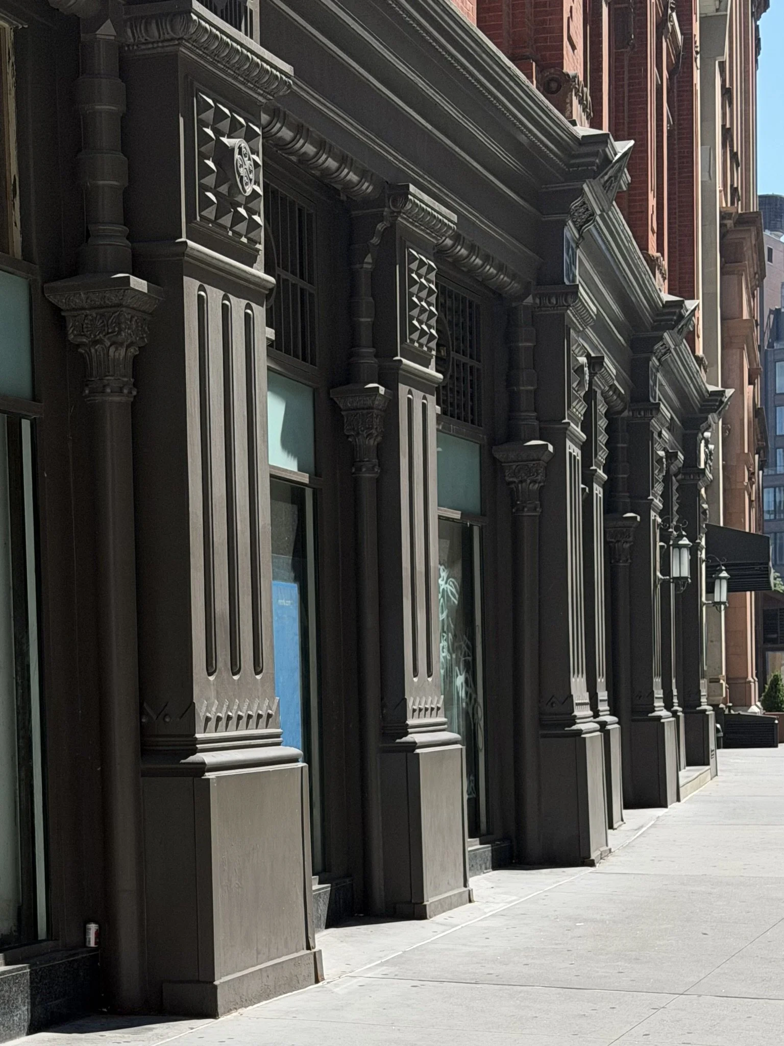 City street with ornate black storefront facades featuring decorative columns and crown molding, buildings with red and beige brick exteriors, and a sidewalk.
