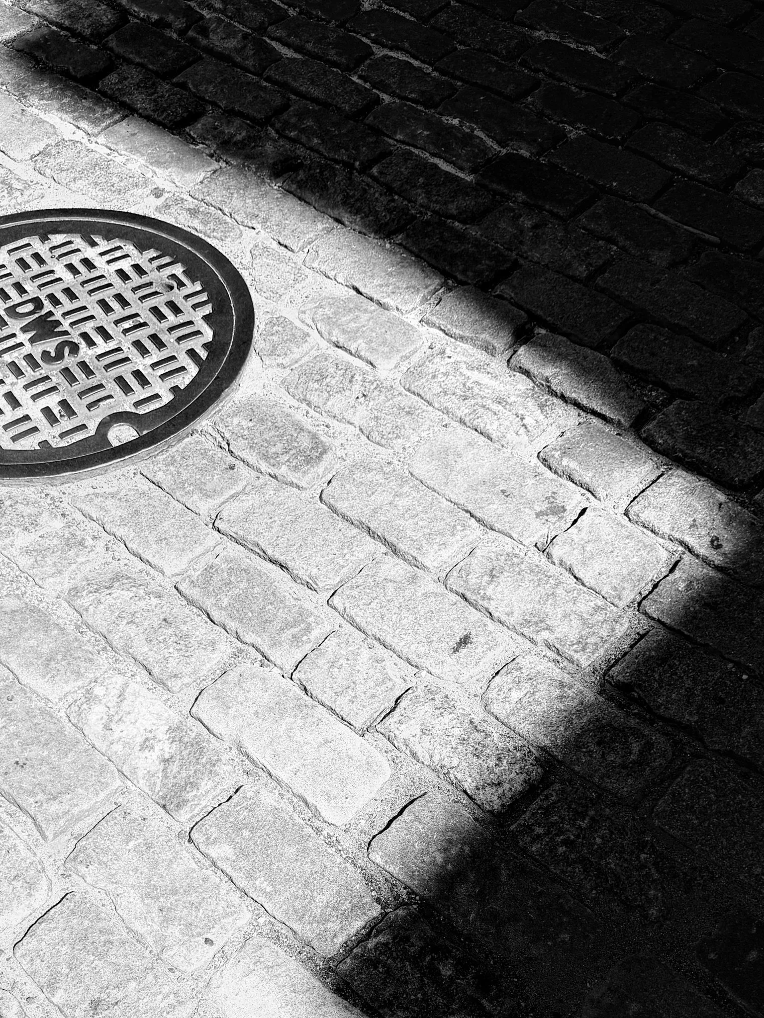 Black and white photo of a round metal water drain cover on a cobblestone street, with a shadow cast over part of the cobblestones.