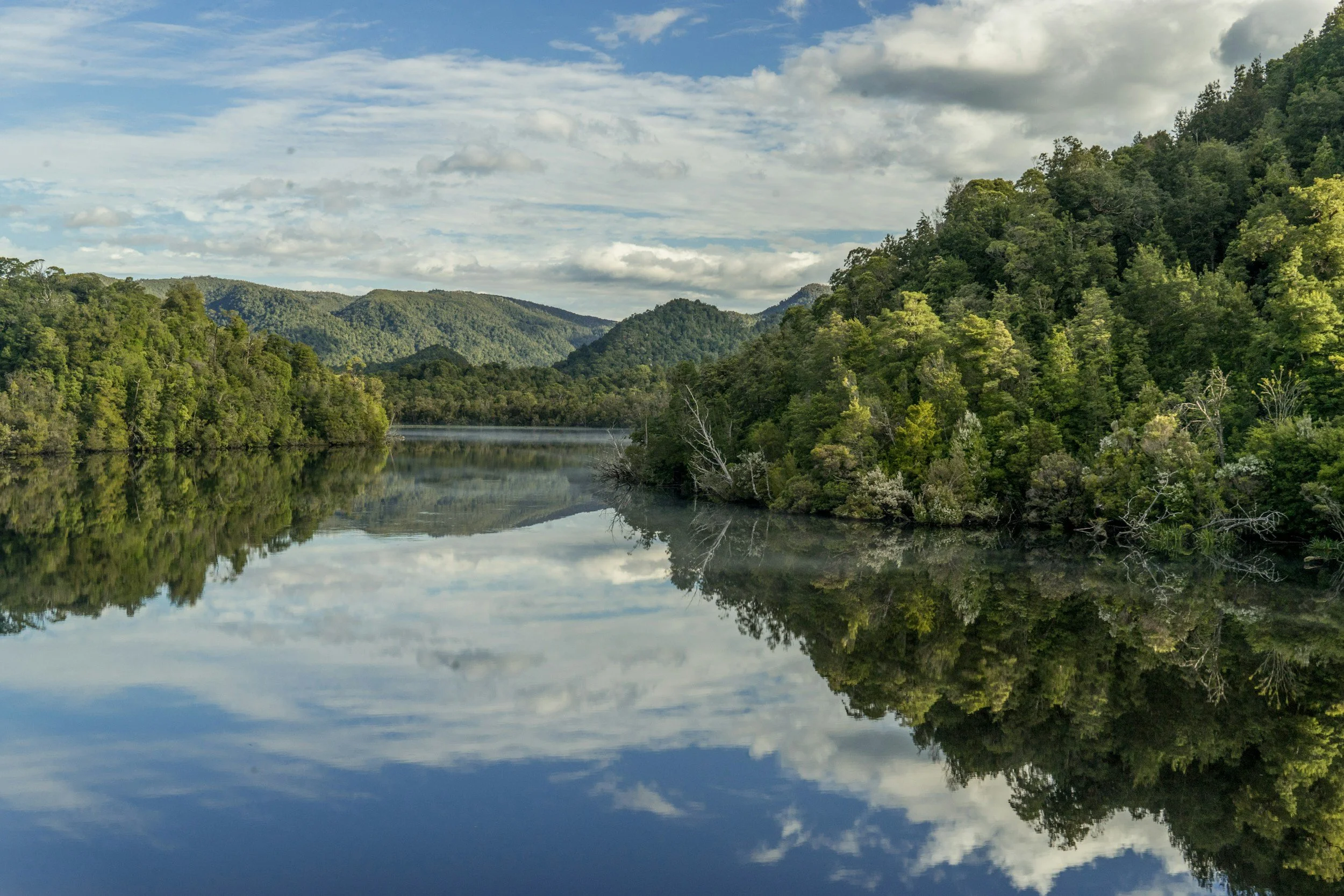 A calm river reflecting green trees on both sides, with a backdrop of rolling hills and a partly cloudy sky.