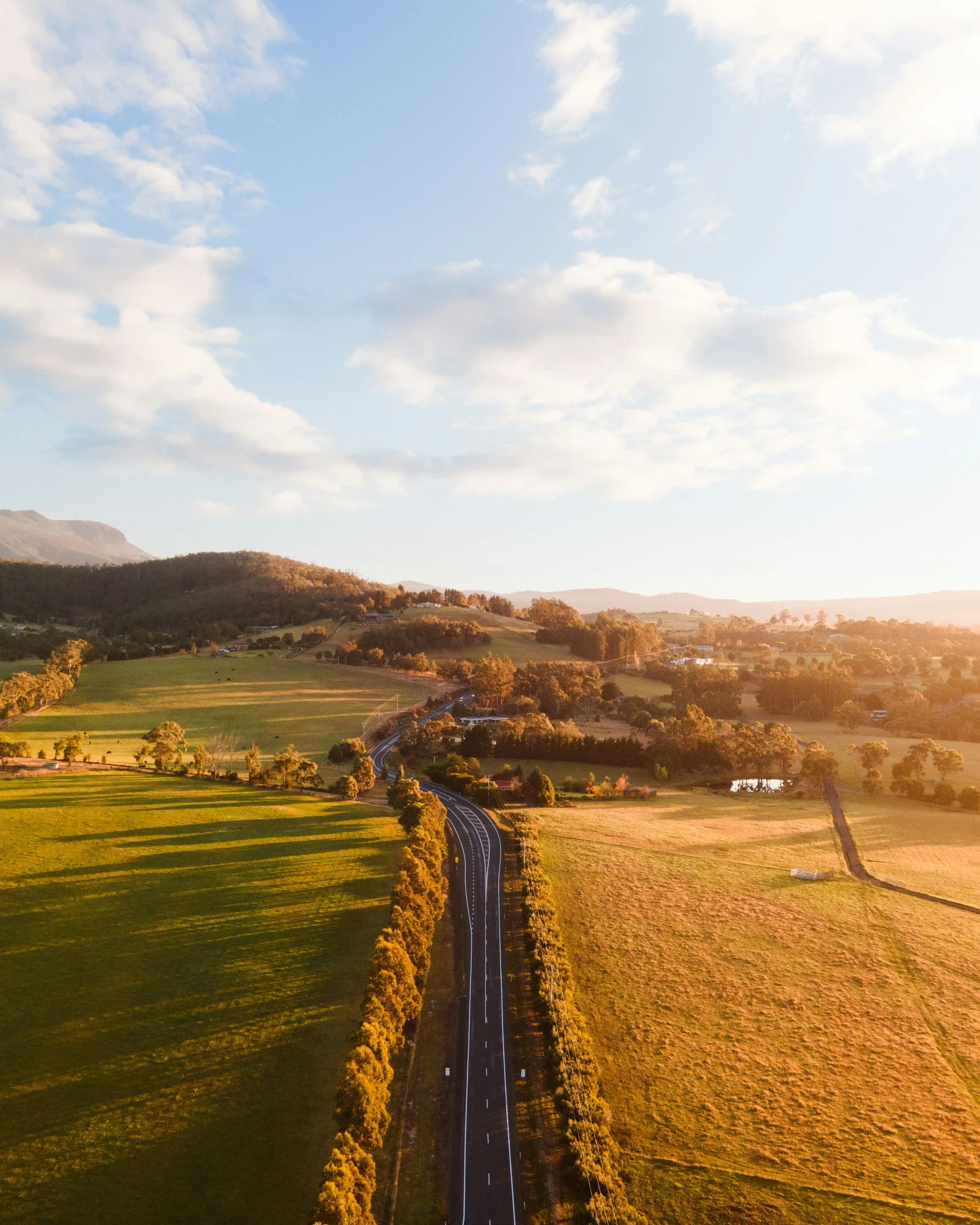 Aerial view of a winding road through green fields and trees, with hills in the background and a partly cloudy sky overhead.