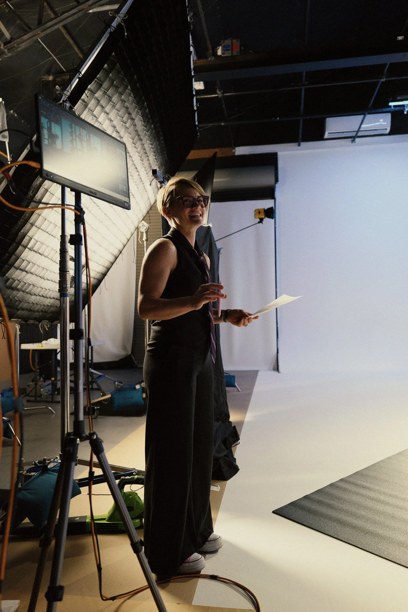 A woman with short blonde hair, glasses, and a sleeveless black top stands in a recording studio, smiling and holding a piece of paper. The studio has a large white screen, lighting equipment, and a textured black wall backdrop.