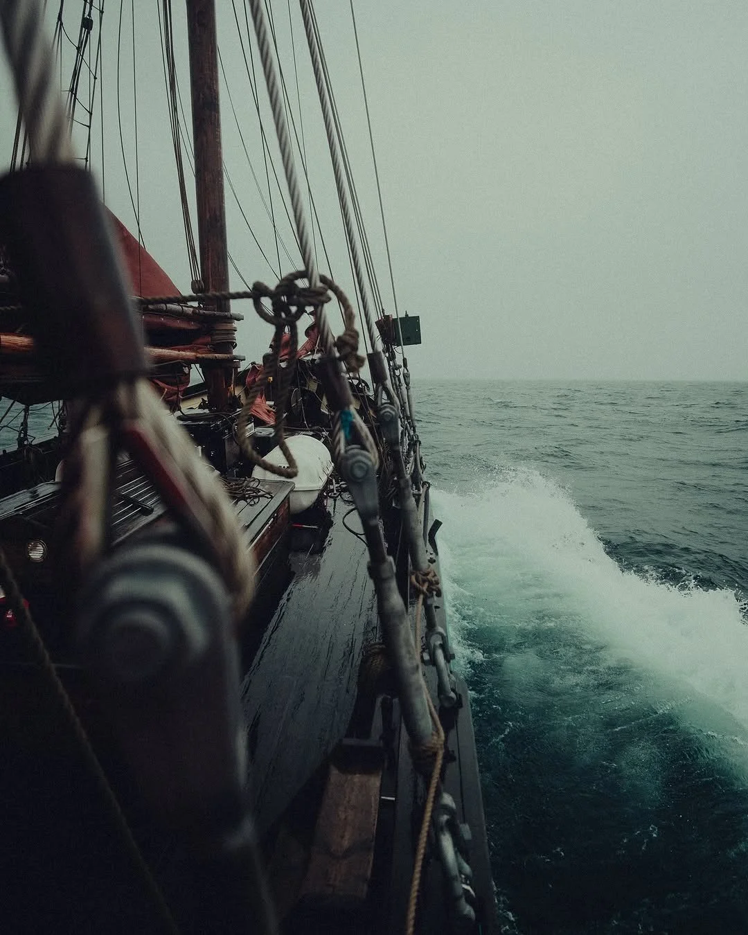 View from a sailboat at sea, showing the boat's wooden deck, rigging, and ropes, with the ocean and horizon in the distance.