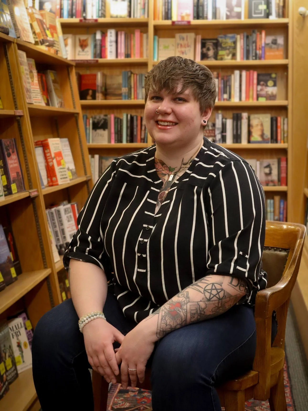 A person with short brown hair, tattoos on their right arm, and piercings on their nose and ears, sitting on a wooden chair in a bookstore surrounded by shelves of books.