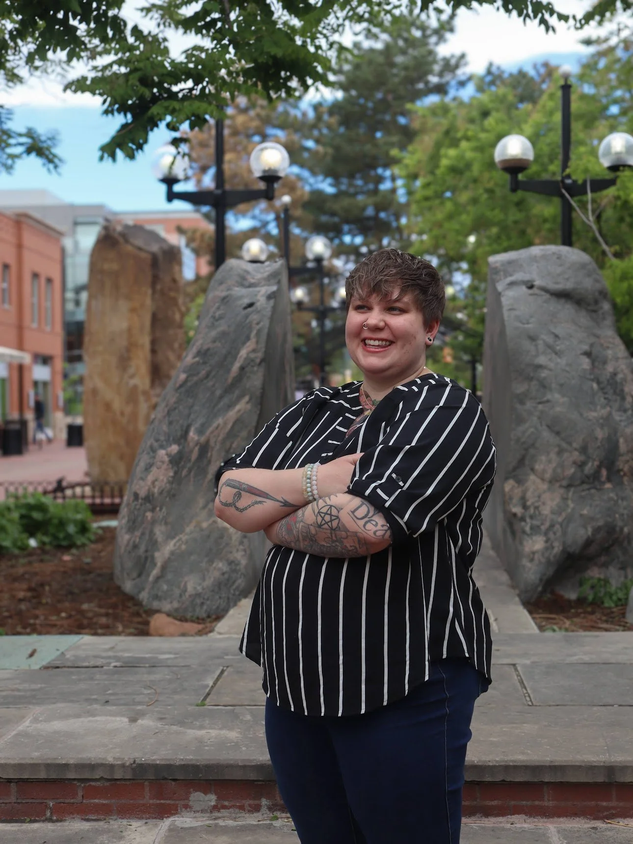 A person with short hair and tattoos on their arms standing outdoors with arms crossed, smiling, in front of large rocks and trees, in an urban park setting.