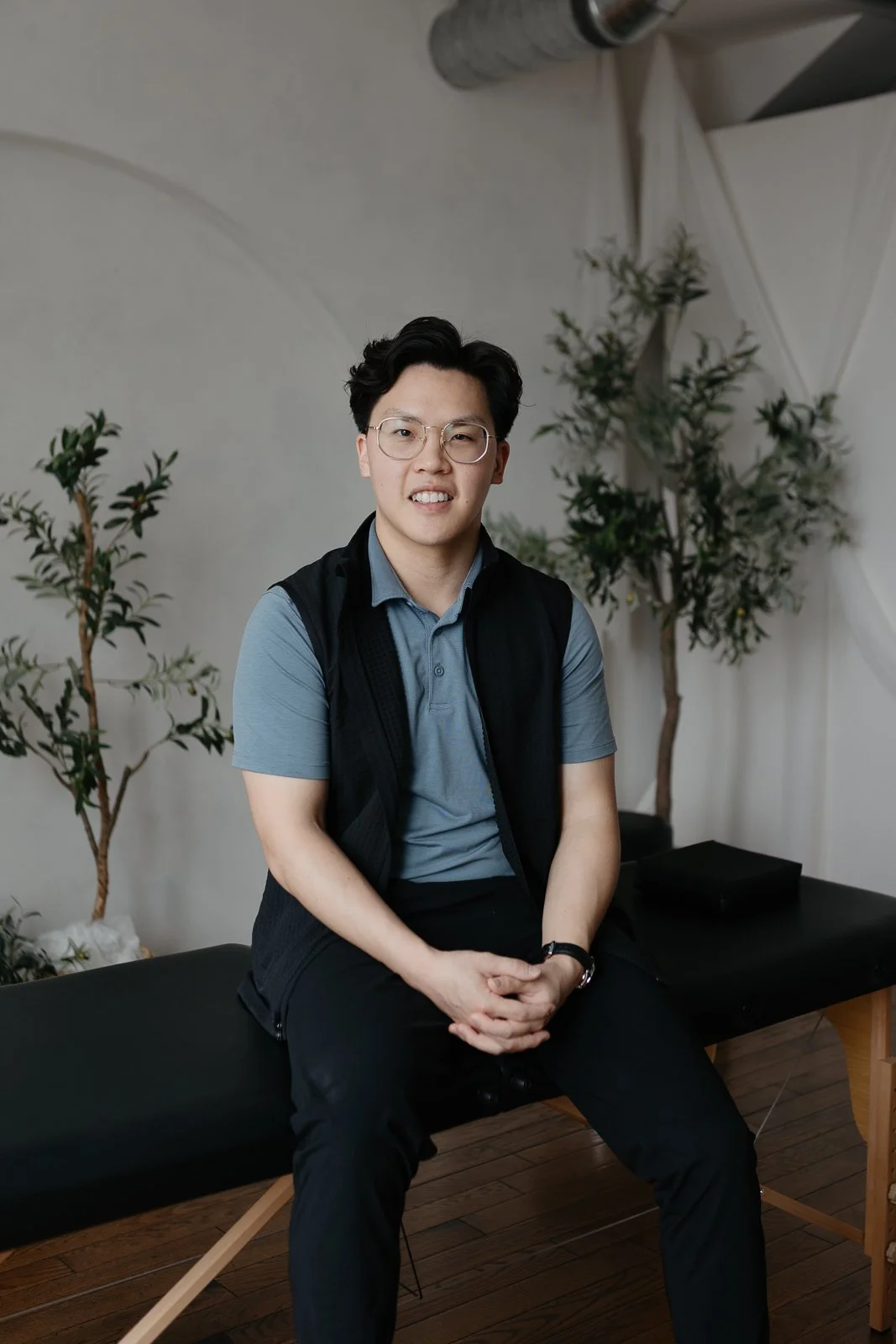 A young man with glasses wearing a blue polo shirt and black pants, sitting on a black bench in front of potted plants and a white wall with draped fabric.