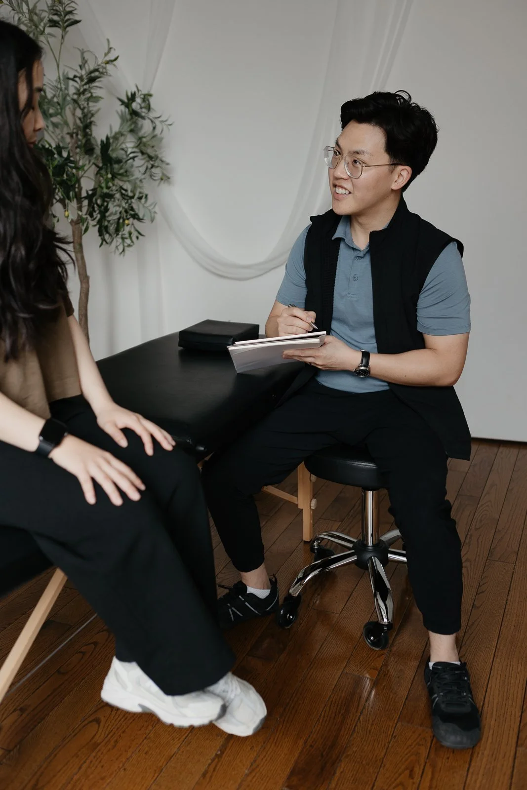 A man with glasses, a black vest, a blue shirt, and black pants sitting on a rolling stool, talking to a woman whose face is not visible, sitting on a chair. The man is holding a notepad and pen, smiling, with a black bag on the table behind him. There is a potted plant and a white curtain in the background.