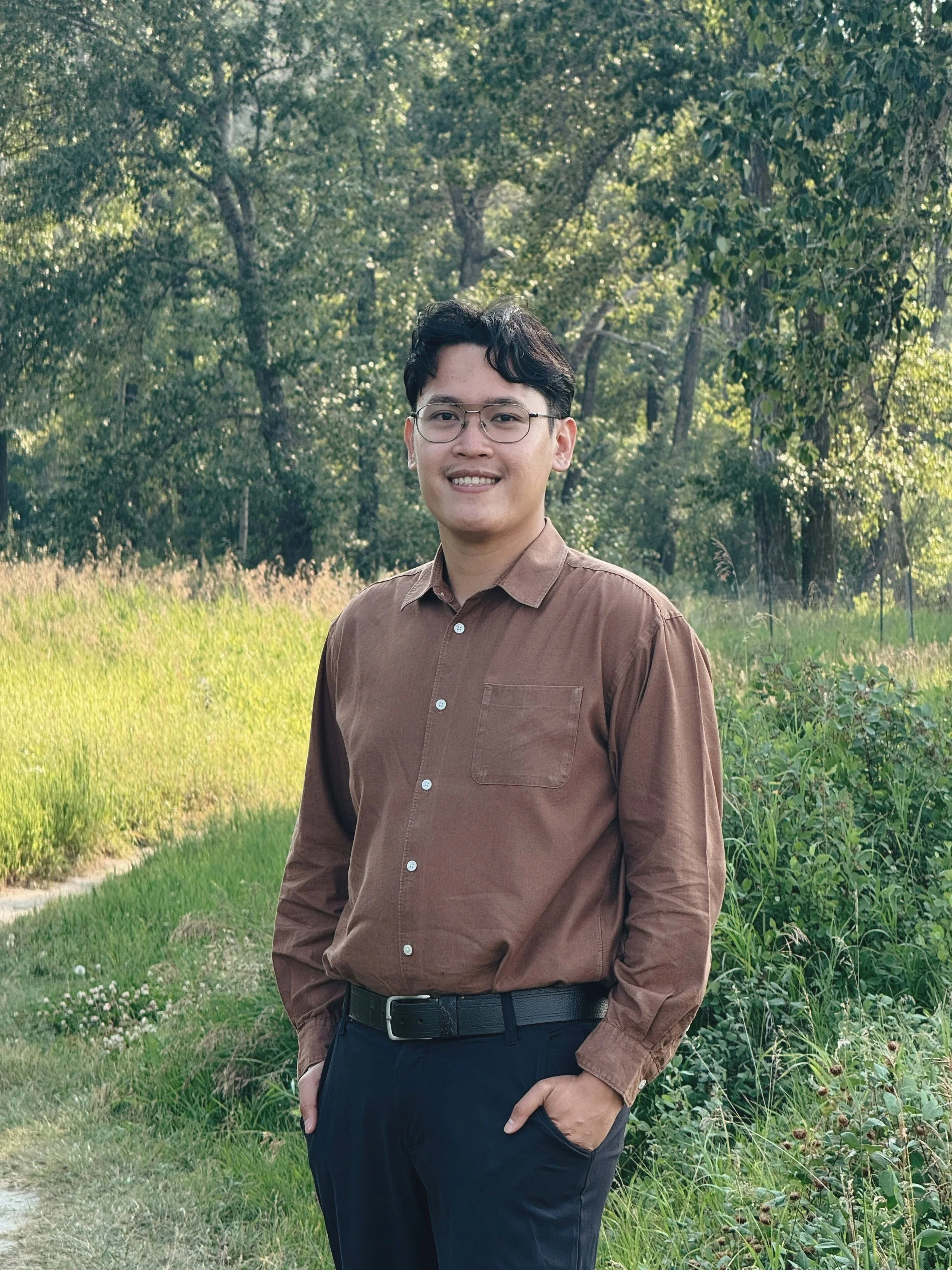 A man with dark hair and glasses wearing a brown button-up shirt and black pants standing outdoors in a grassy area with trees in the background.