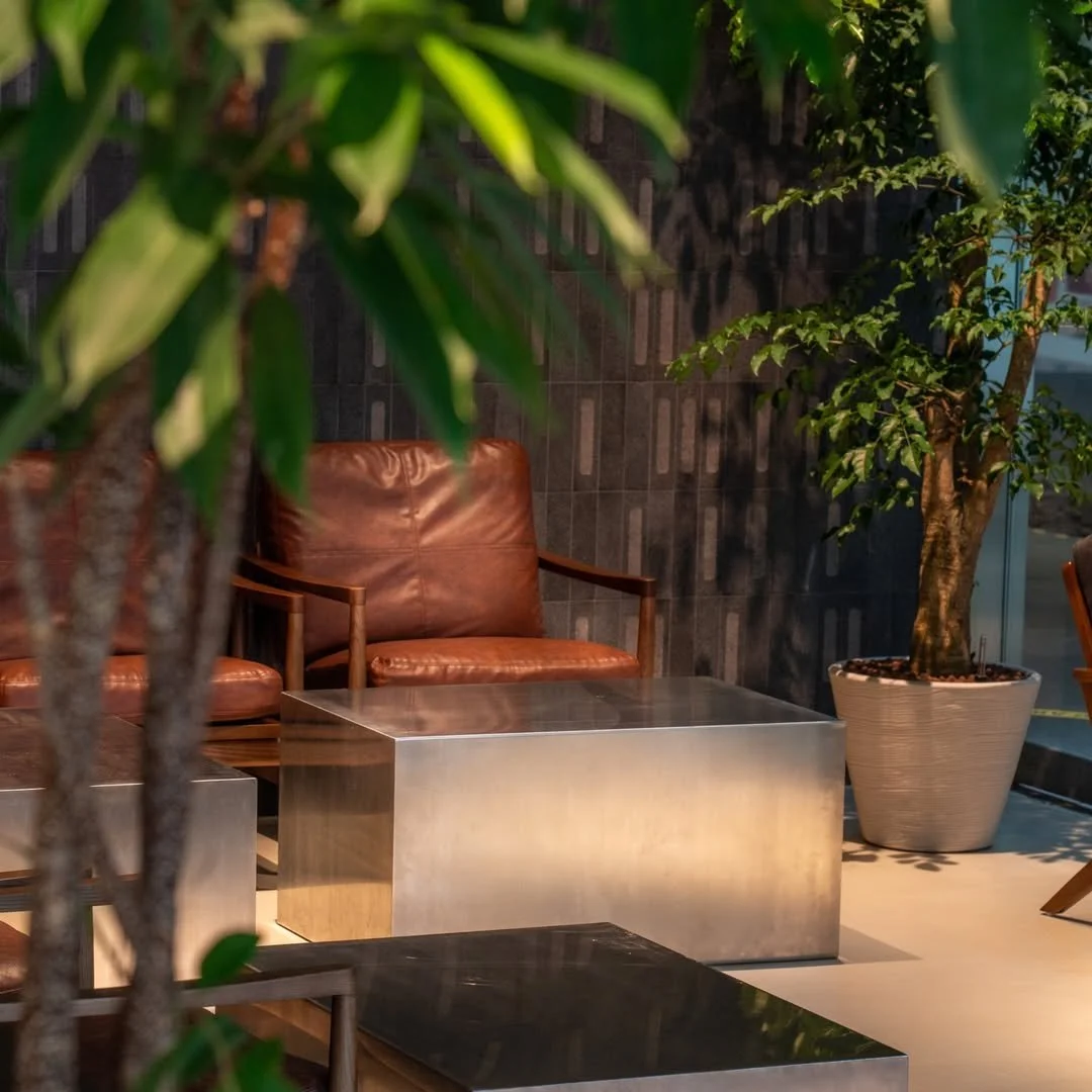 Indoor seating area with brown leather chairs, metallic tables, and potted trees, with a dark wall and large window in the background.