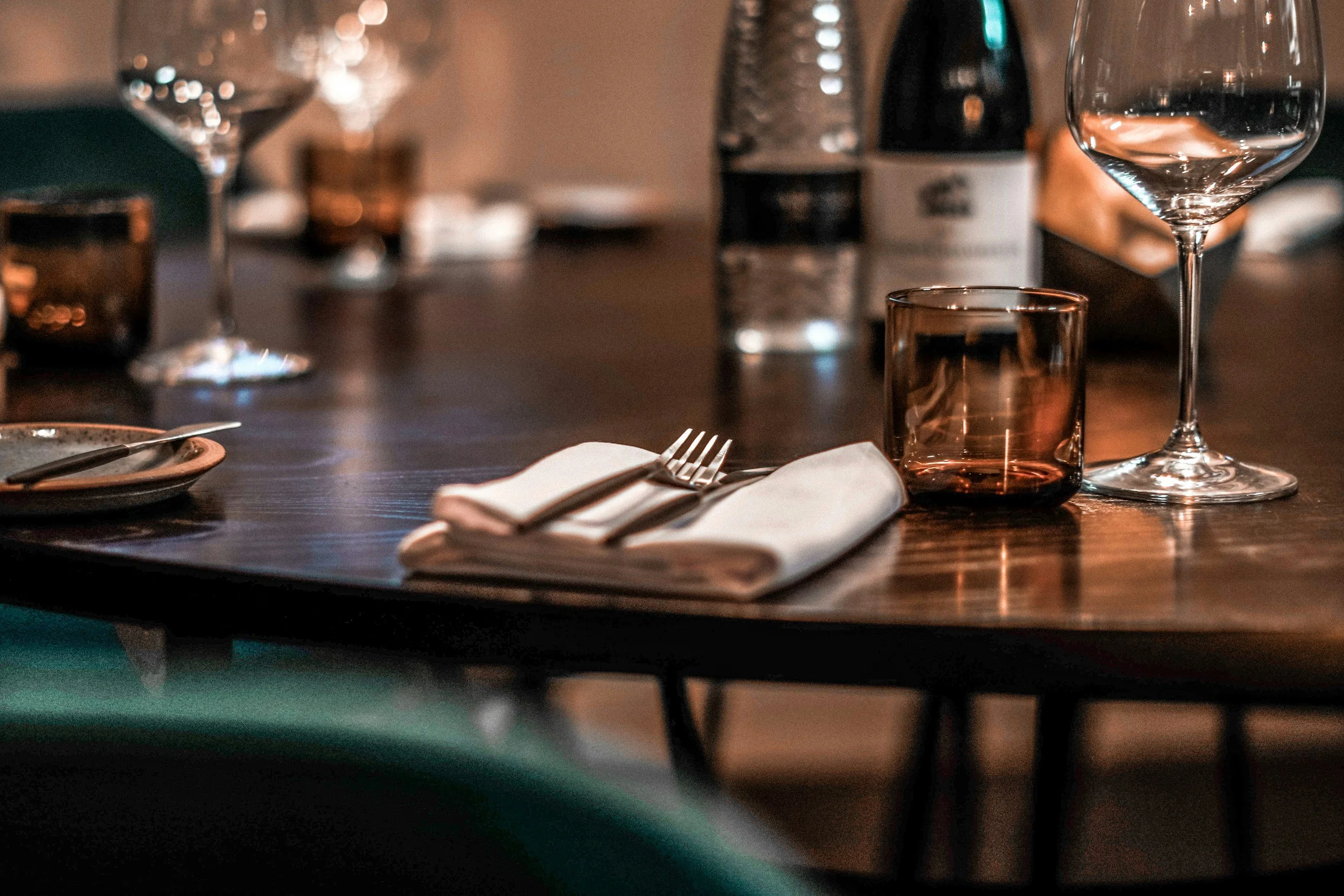Dining table set with wine glasses, a small plate, fork, knife, napkin, and a shot glass, with blurred bottles in the background.