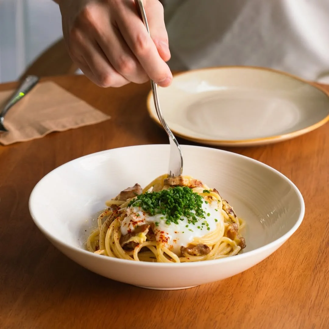 A person is garnishing a plate of pasta with chopped green herbs and a poached egg on top.