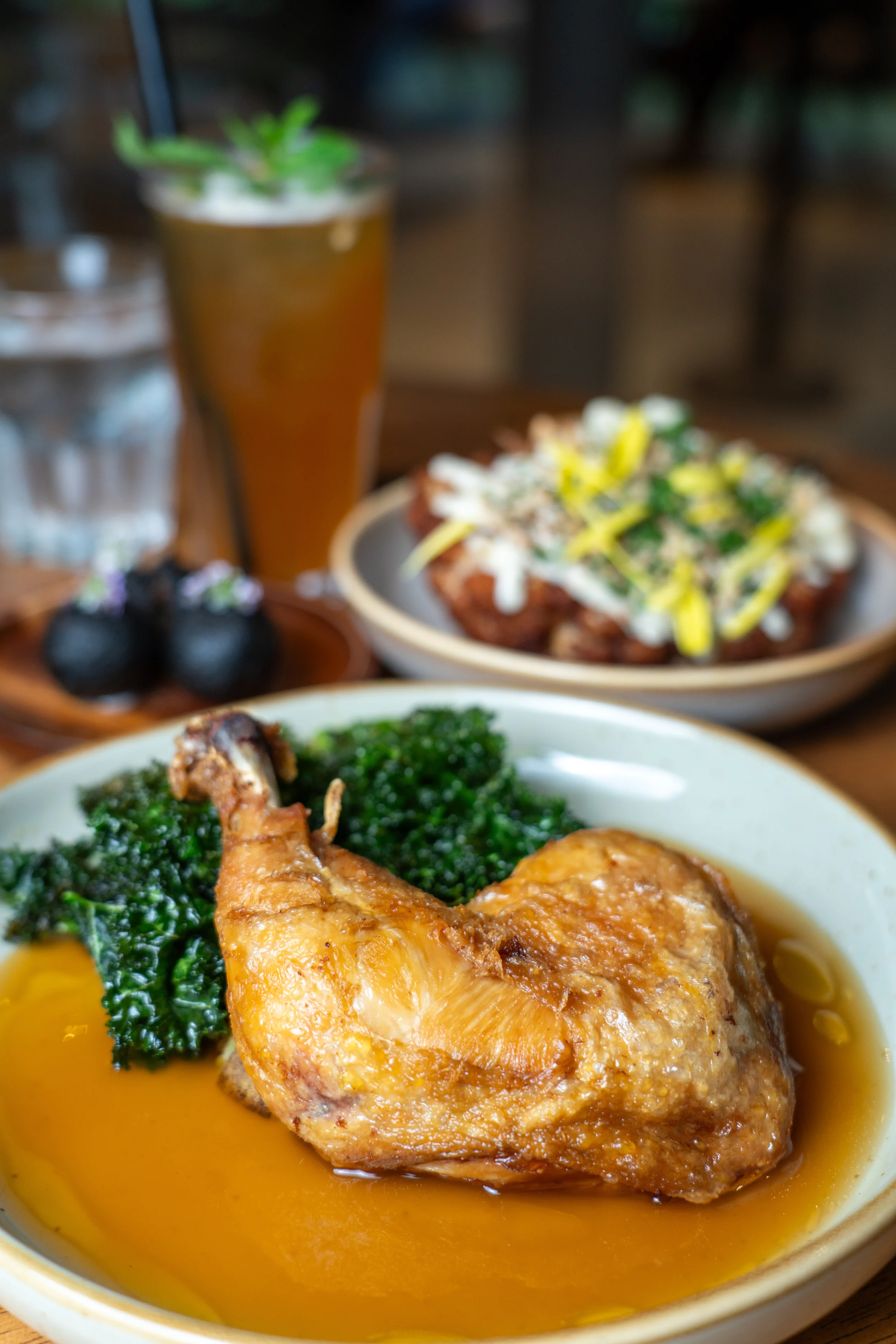 Close-up of a bowl with a roasted chicken leg, wilted greens, and a flavorful broth. In the background, a plate with a colorful salad topped with shredded cheese, and a tall glass of iced tea garnished with a sprig of mint, alongside a glass of water.