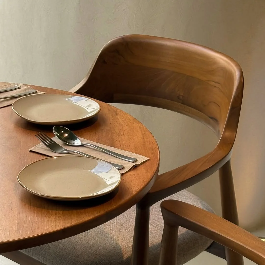 A wooden dining table with two beige plates, two forks, two spoons on matching napkins, and a wooden chair with a curved backrest.