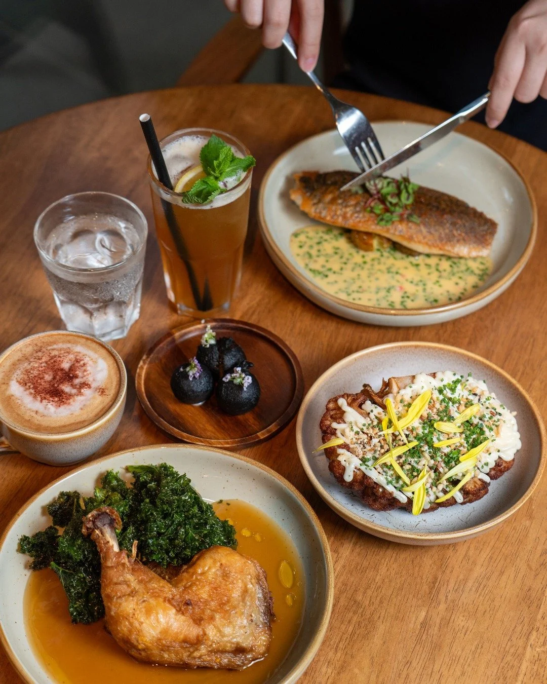 A table with various dishes including a plate of red fish with sauce, a bowl of fried chicken with greens, a plate of topped fried food, a glass of mojito, a glass of water, a cup of coffee with cinnamon, and small dessert balls garnished with flower