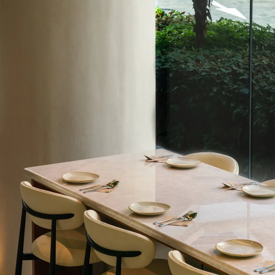 A dining table set with plates, forks, and spoons near a large window with greenery outside.