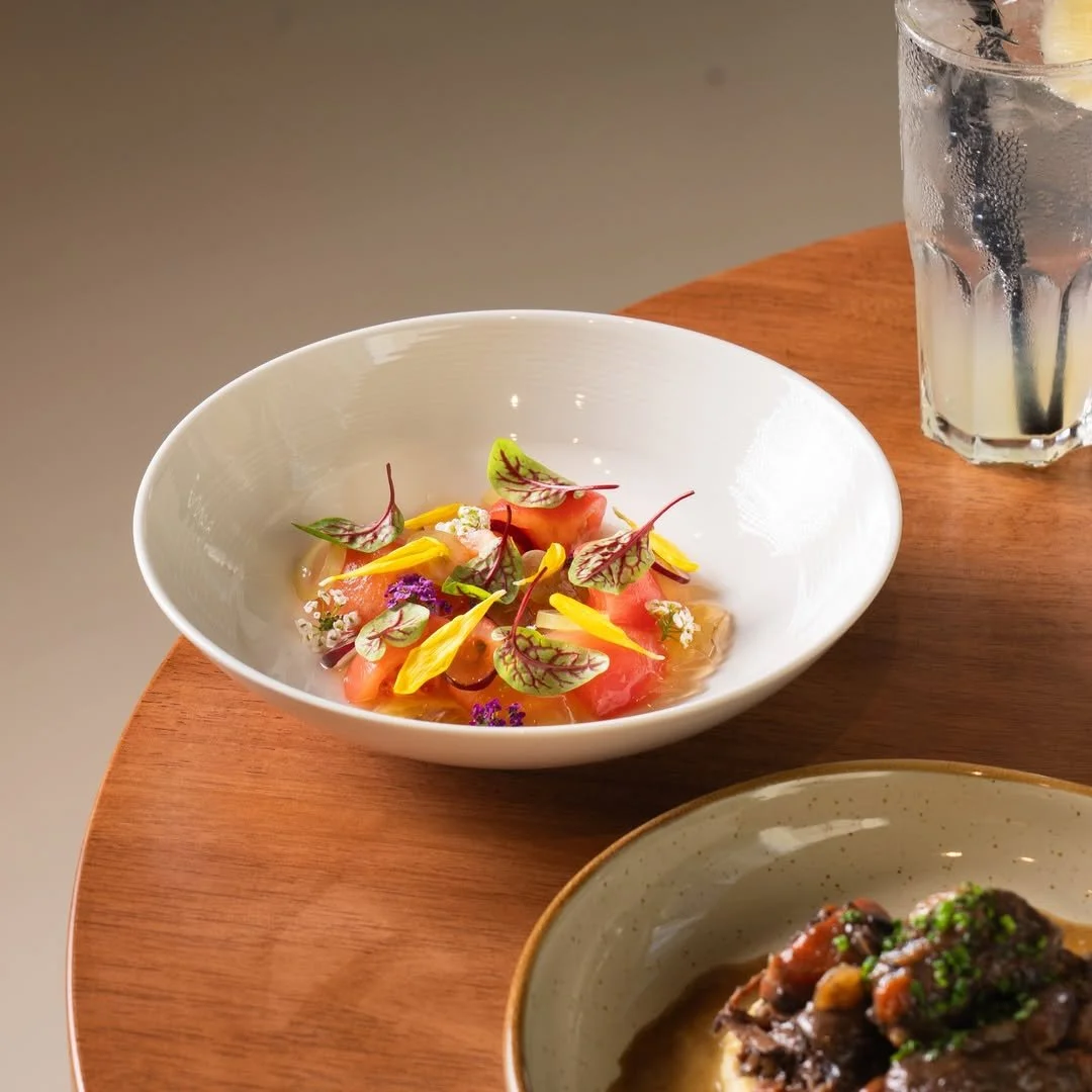 A white bowl of colorful salad with edible flowers and microgreens, a glass of lemonade with ice and lemon slices, and a bowl of cooked meat with herbs on a wooden table.