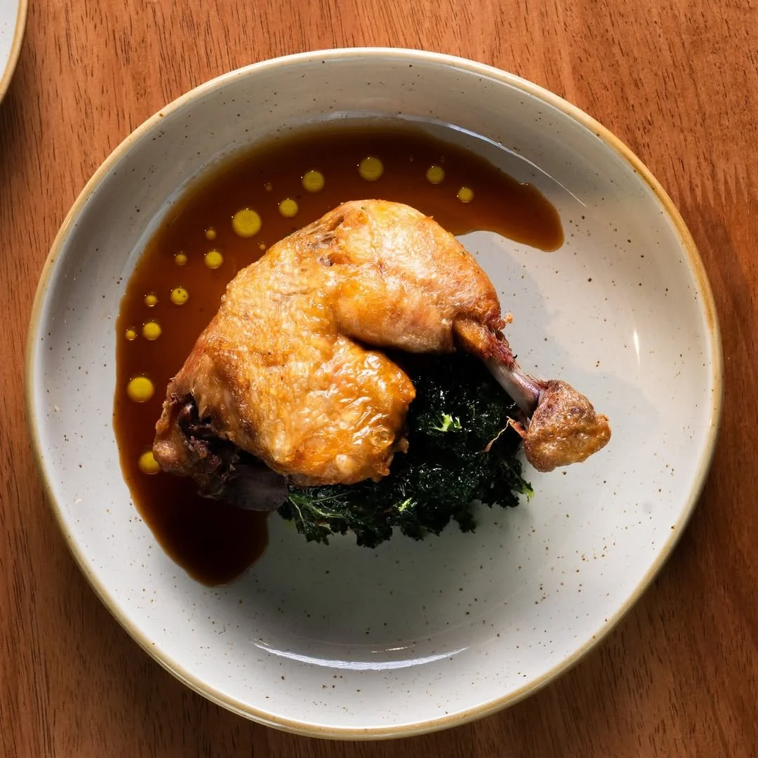 Cooked chicken thigh on a bed of greens with brown sauce and yellow oil droplets in a bowl on a wooden table.