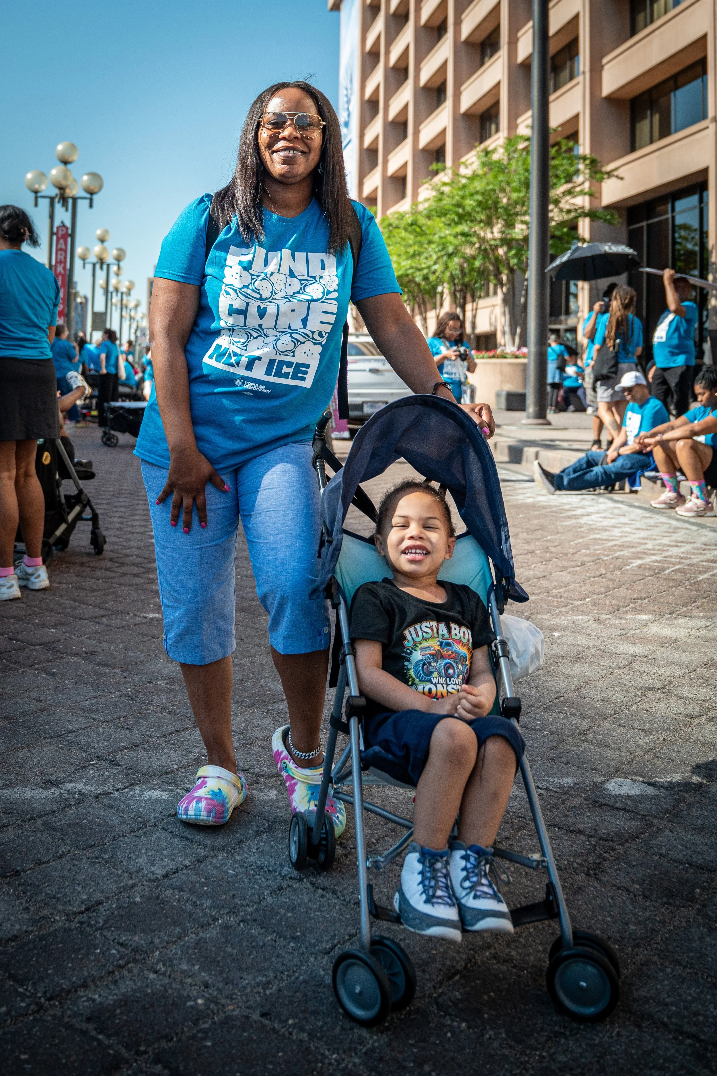 A woman standing on a city street, pushing a smiling young boy in a stroller. The woman is wearing colorful sneakers and sunglasses, while the boy is wearing a black T-shirt and colorful sneakers. Several people are sitting and standing in the background, some holding umbrellas, against buildings and trees on a sunny day.