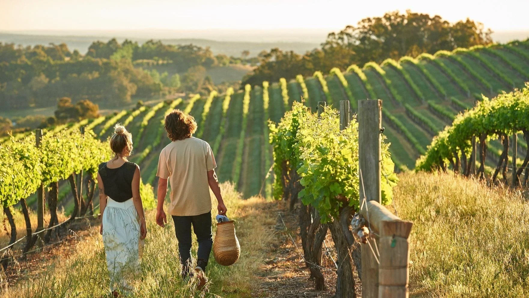 Two people, a man and a woman, walking through a vineyard during sunset. The woman has blonde hair tied up, wearing a black sleeveless top and white skirt. The man has curly brown hair, wearing a beige t-shirt, dark pants, and holding a woven basket. Rows of grapevines are on either side, with rolling hills in the background.