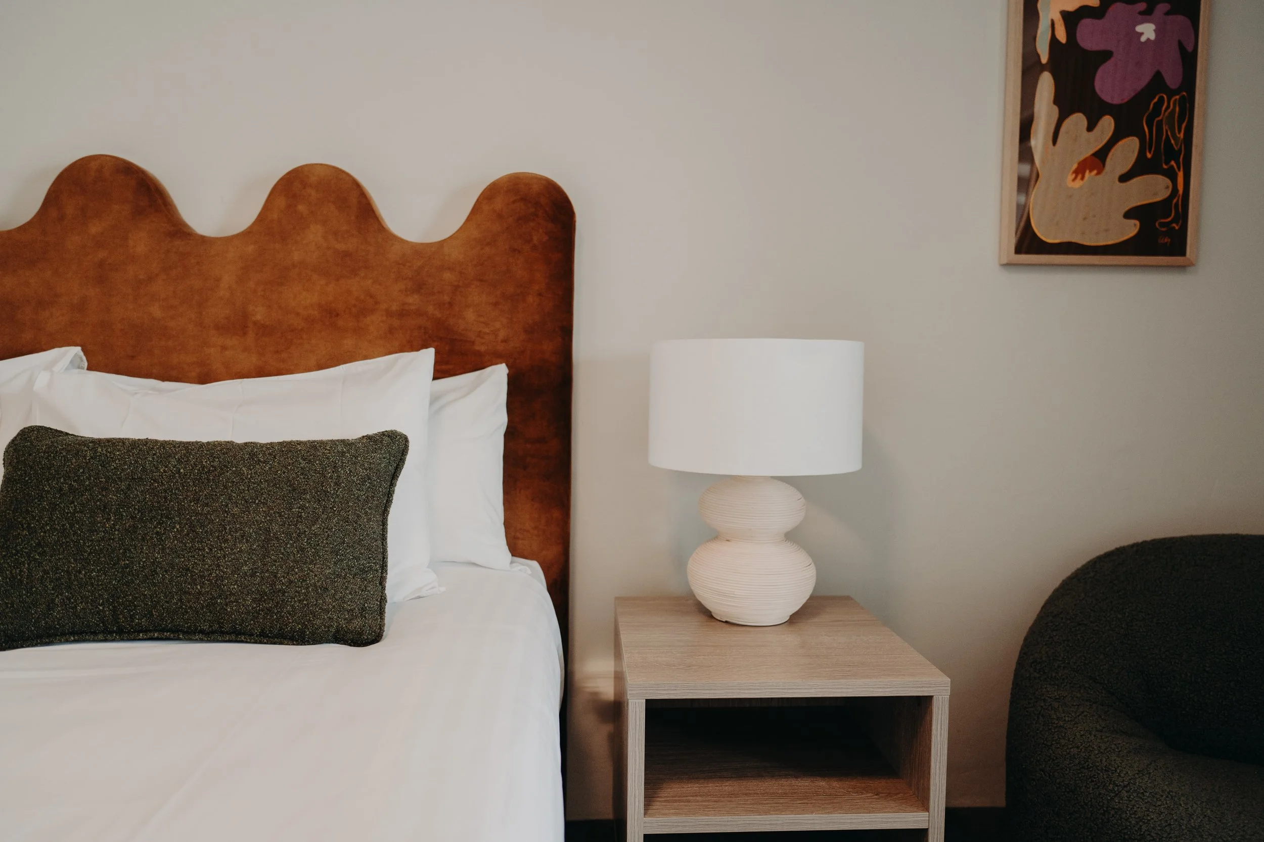 A bedroom with a wooden headboard, white bedding, pillows, a bedside table, a white table lamp, and a piece of modern wall art with abstract shapes.