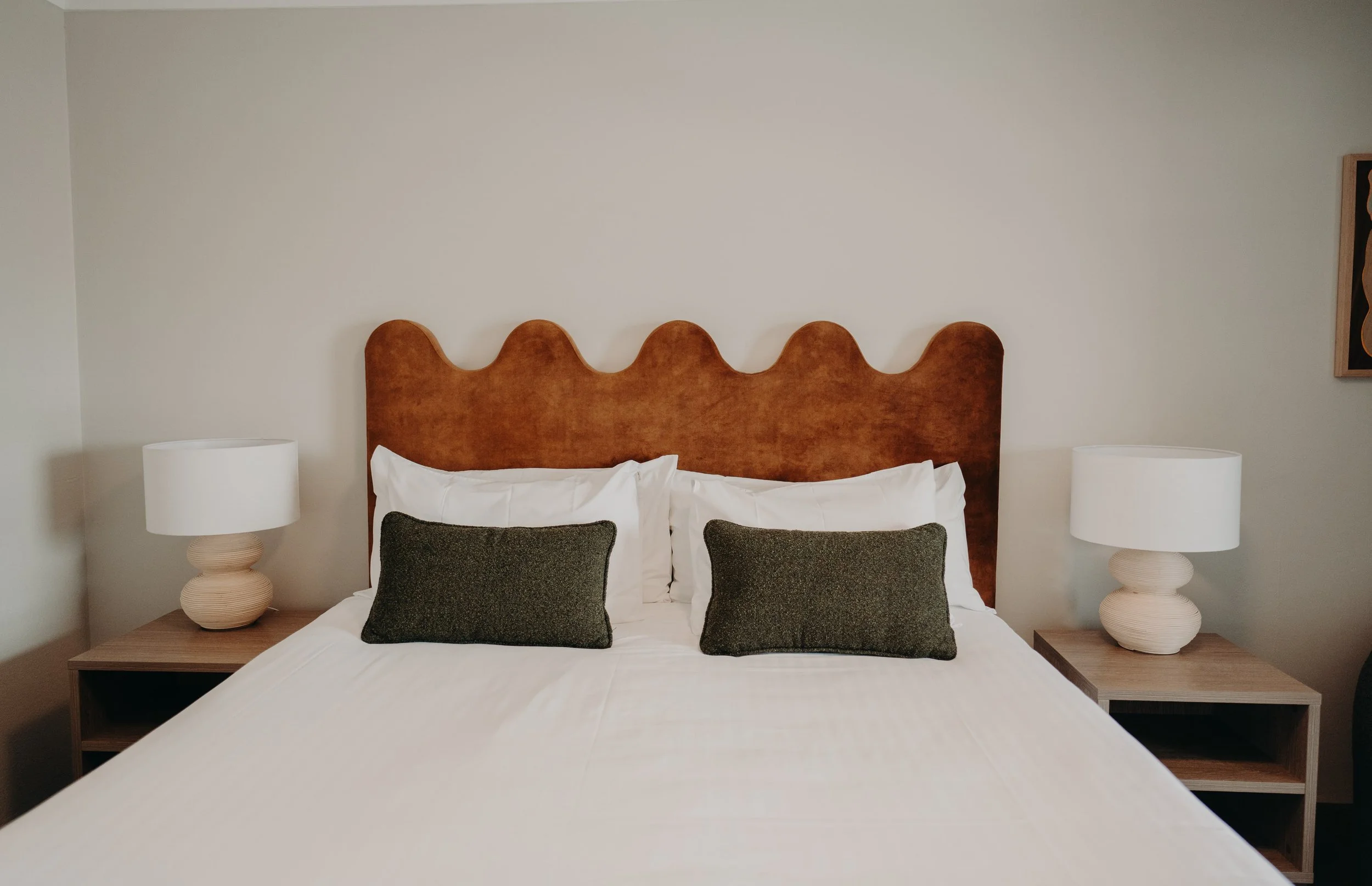 A neatly made bed with white bedding, dark textured pillows, and a wooden headboard with a wavy design. On either side, there are matching wooden nightstands with modern table lamps featuring sculpted, rounded bases and white shades. The background is a plain light-colored wall with minimal decor.