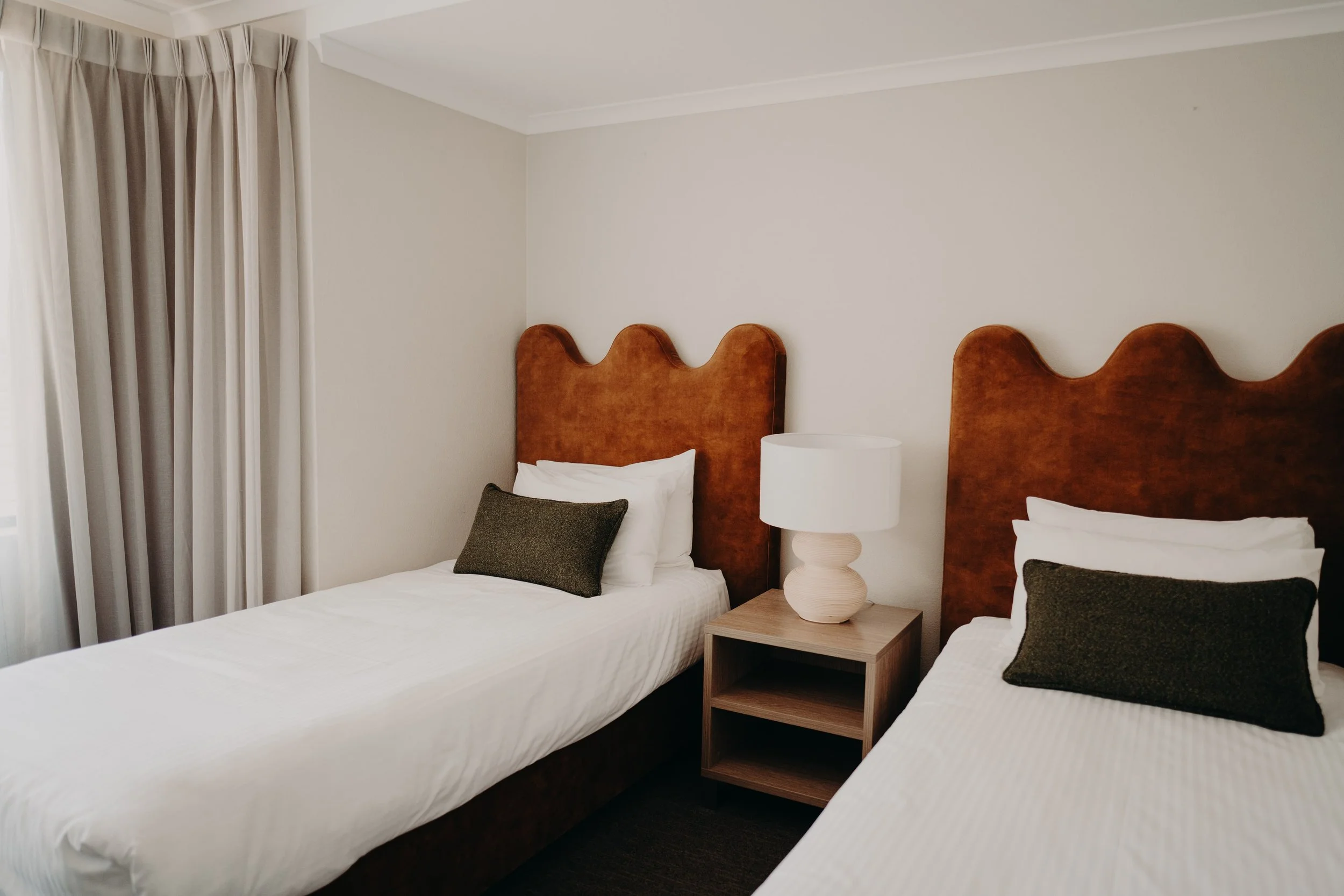 Hotel room with two beds, each with a brown upholstered headboard, white bedding, and dark gray pillows. A small wooden nightstand with a modern beige lamp is between the beds. White curtains cover a window in the corner of the room, and the walls are painted light beige.