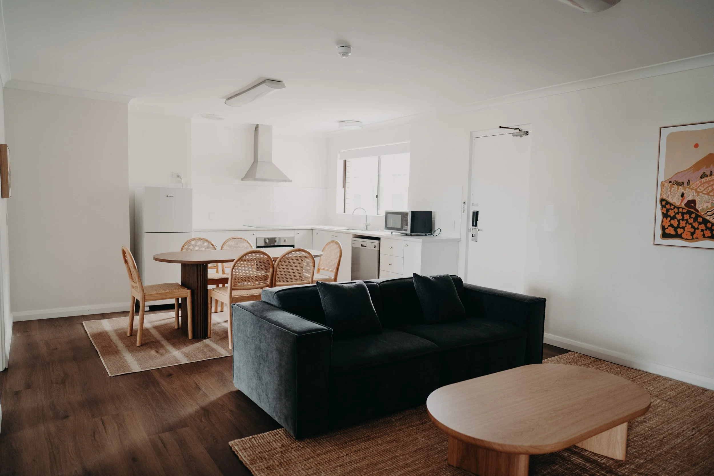 Open-concept living room and kitchen with white walls, wooden flooring, a black sofa, a wooden dining table with six chairs, a white refrigerator, microwave, dishwasher, artwork on the wall, and natural light from the window.