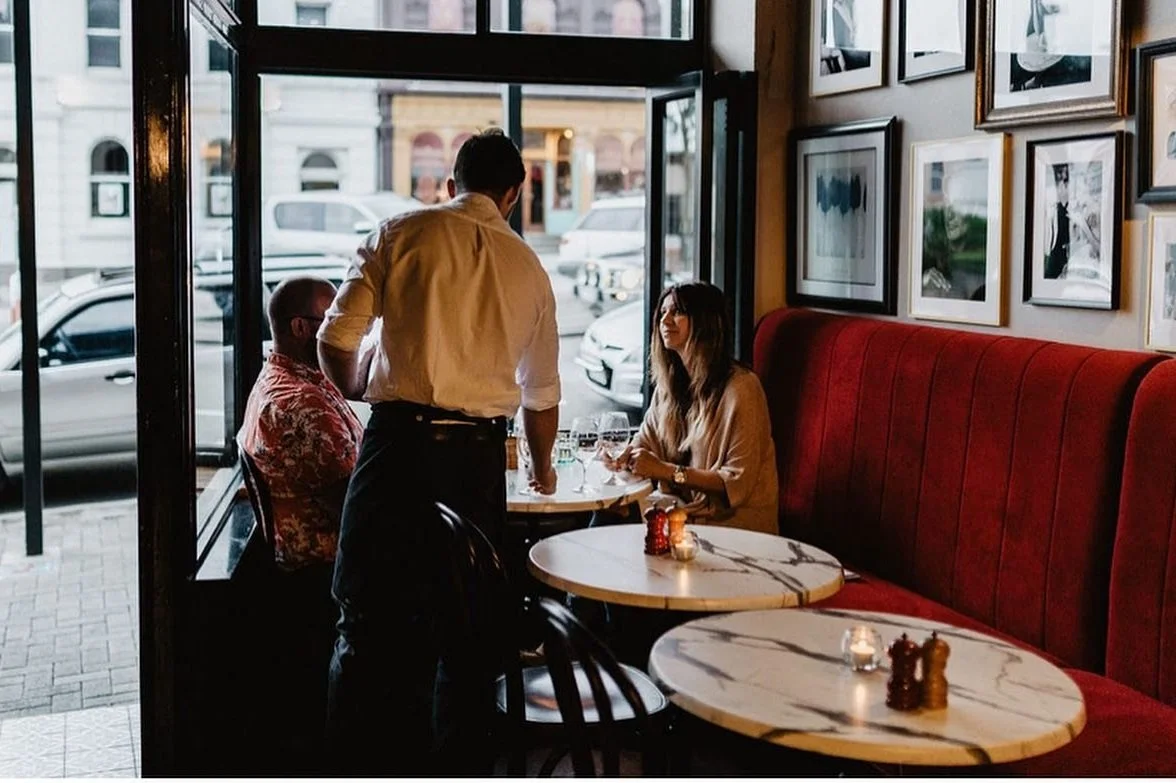 Interior of a restaurant with a man standing and two customers sitting at a table near the window. The woman is seated on a red bench, and there are picture frames on the wall. Outside the window, cars are visible.