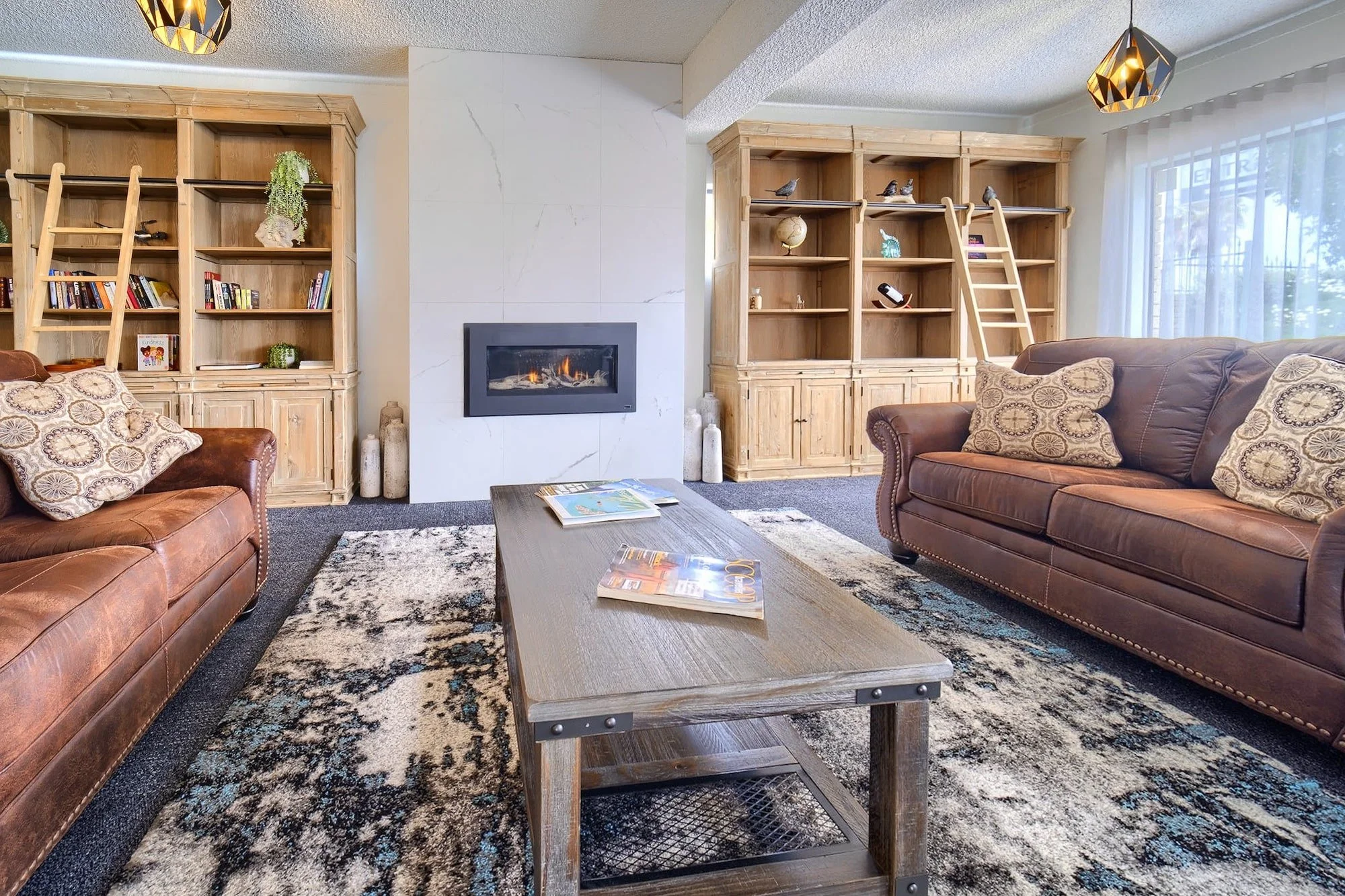 Living room with brown leather sofas, wooden bookshelves, a fireplace, a coffee table with magazines, a patterned rug, and large windows with sheer curtains.