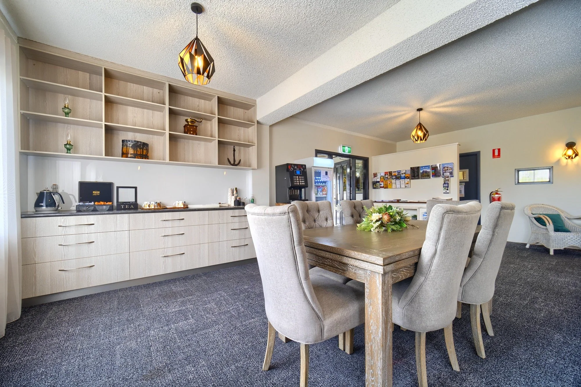 Interior of a lounge or dining area with a wooden table and beige upholstered chairs, a built-in cabinet with decorative items, a coffee machine, and a vending machine, with ceiling lights and a small window.