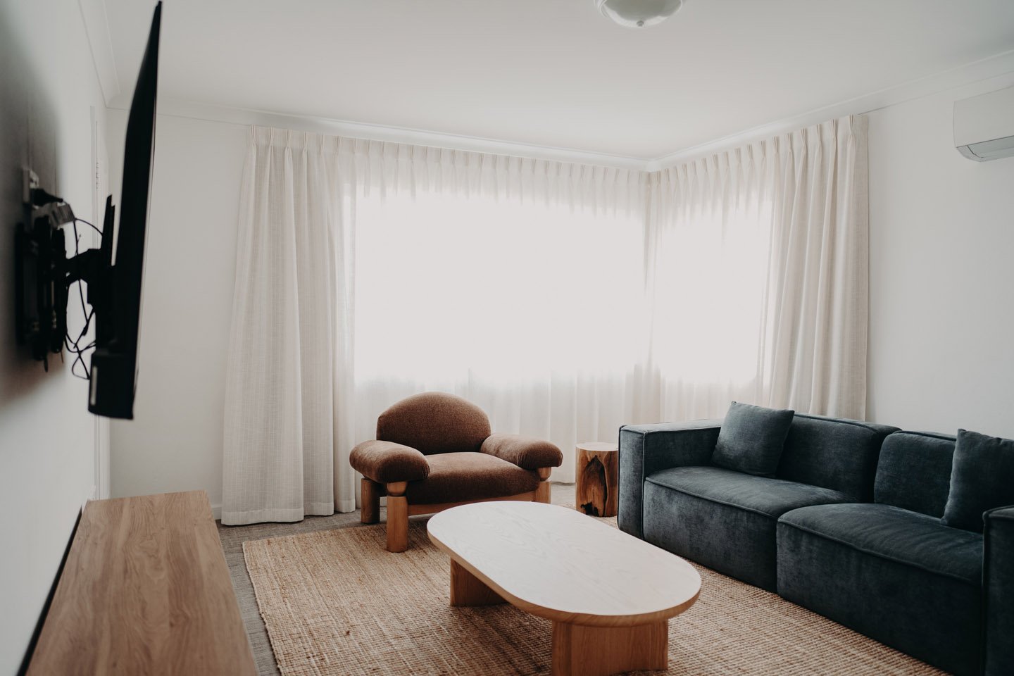 Living room with white walls, large window with cream curtains, a blue velvet sofa with pillows, a brown armchair, a wooden coffee table, a side table, and an air conditioning unit.