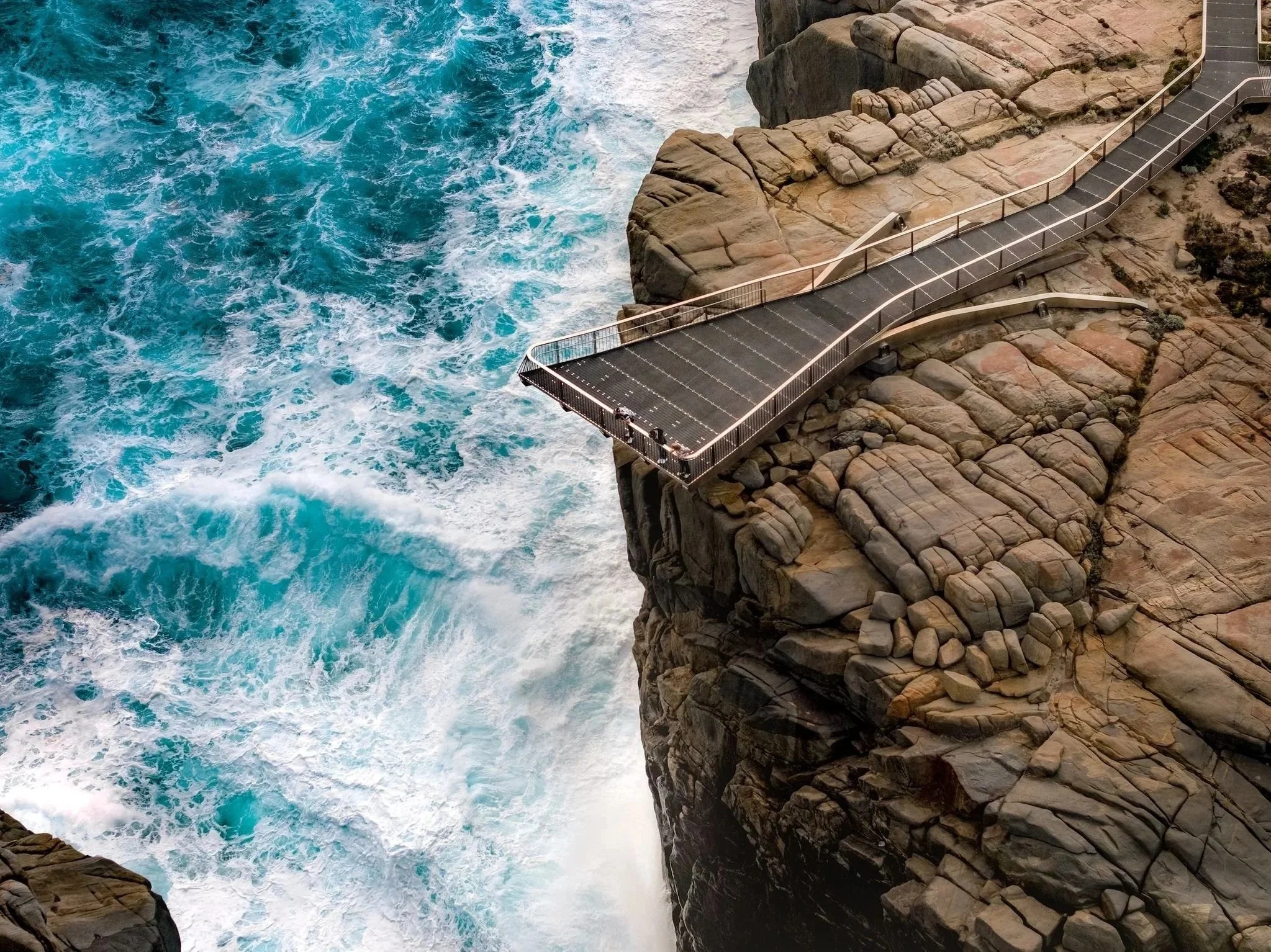 A rocky cliffside with a pathway and railing overlooking the ocean, with waves crashing against the rocks.