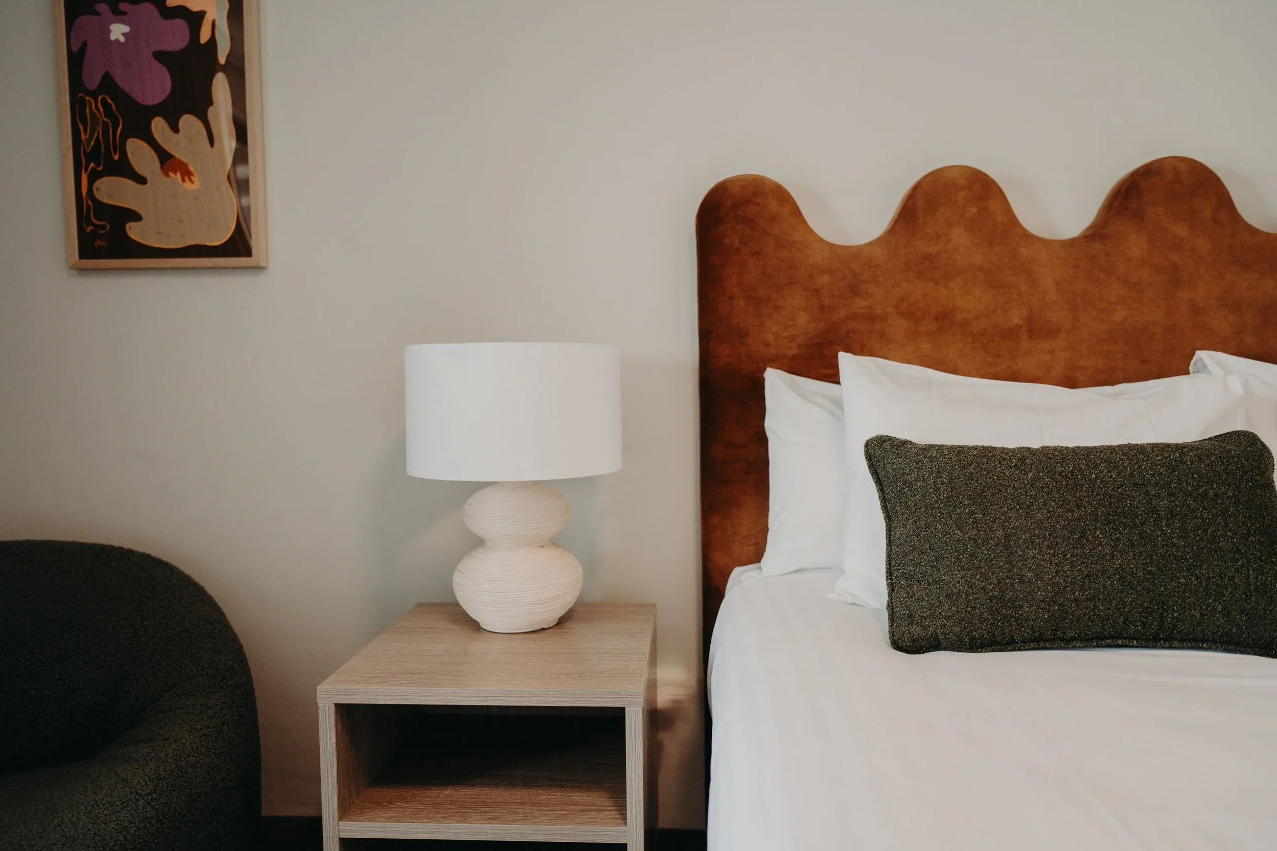 Interior bedroom with a wooden headboard, white pillows, a textured black pillow, a white table lamp, a small wooden side table, and a framed abstract art piece on the wall.