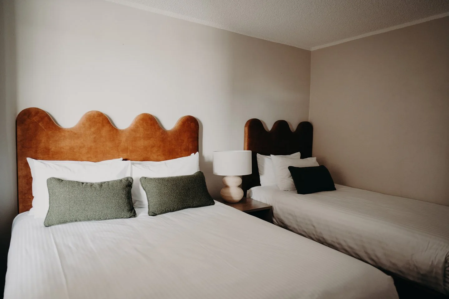 Two beds with brown headboards, white bedding, and assorted pillows, situated in a hotel room with beige walls and a nightstand with a lamp between the beds.