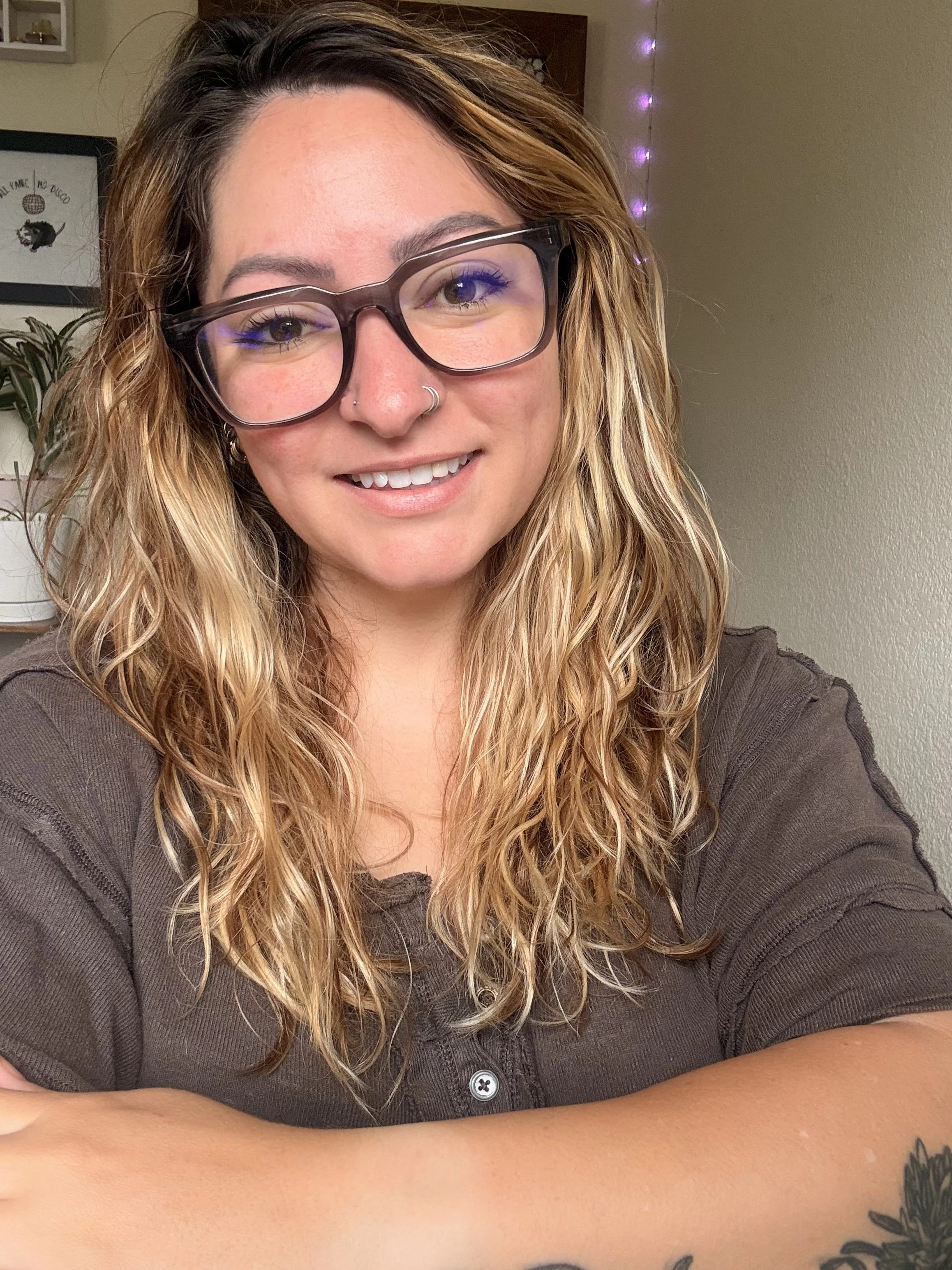 Smiling woman with wavy blonde hair, dark-framed glasses, and nose piercings posing indoors.
