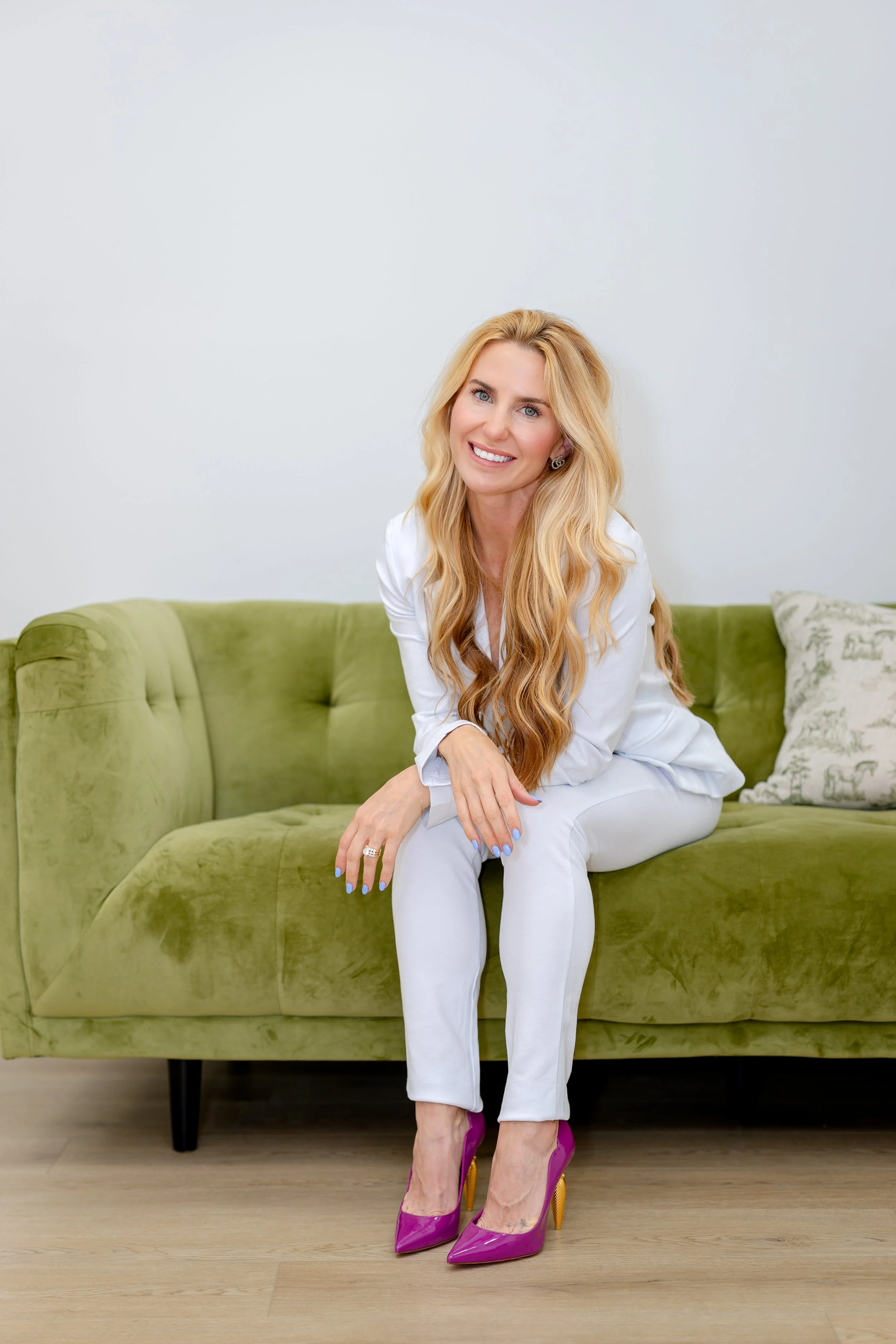 A woman with long blonde hair, blue eyes, and fair skin is sitting on a green sofa in front of a plain white wall. She is smiling and wearing a white outfit and bright pink high-heeled shoes. She has a ring and earrings, and cowboy-style manicured nails.