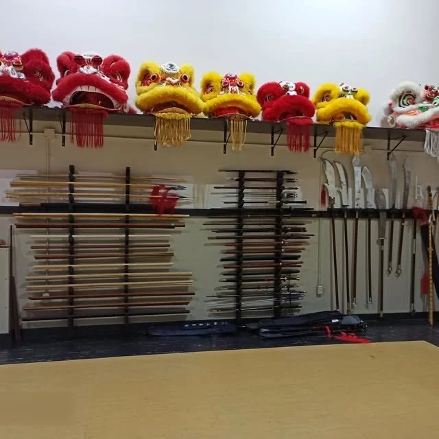 Row of colorful lion dance costumes on a shelf above martial arts weapons stored on wall racks.