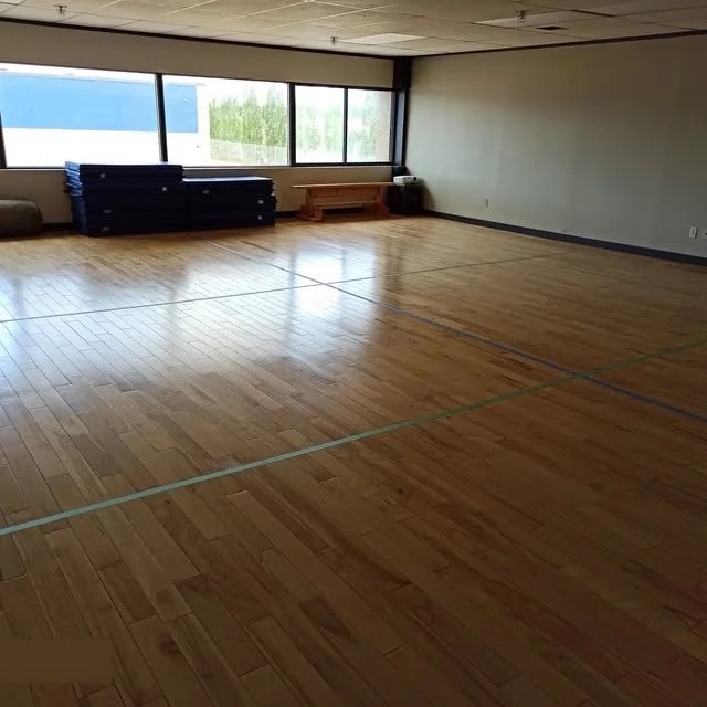Empty indoor gymnasium with wooden floor, large windows, and stacked gym mats against the wall.