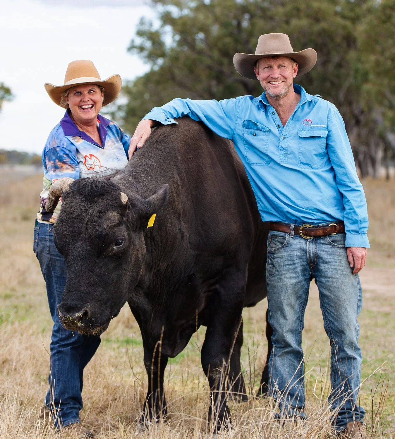 Two people, a woman and a man, standing outdoors with a black calf. The woman wears a straw hat and colorful cowboy shirt, smiling; the man wears a beige cowboy hat and light blue shirt, smiling and resting his arm on the calf. The setting appears to be a rural or farm area with trees and grass.