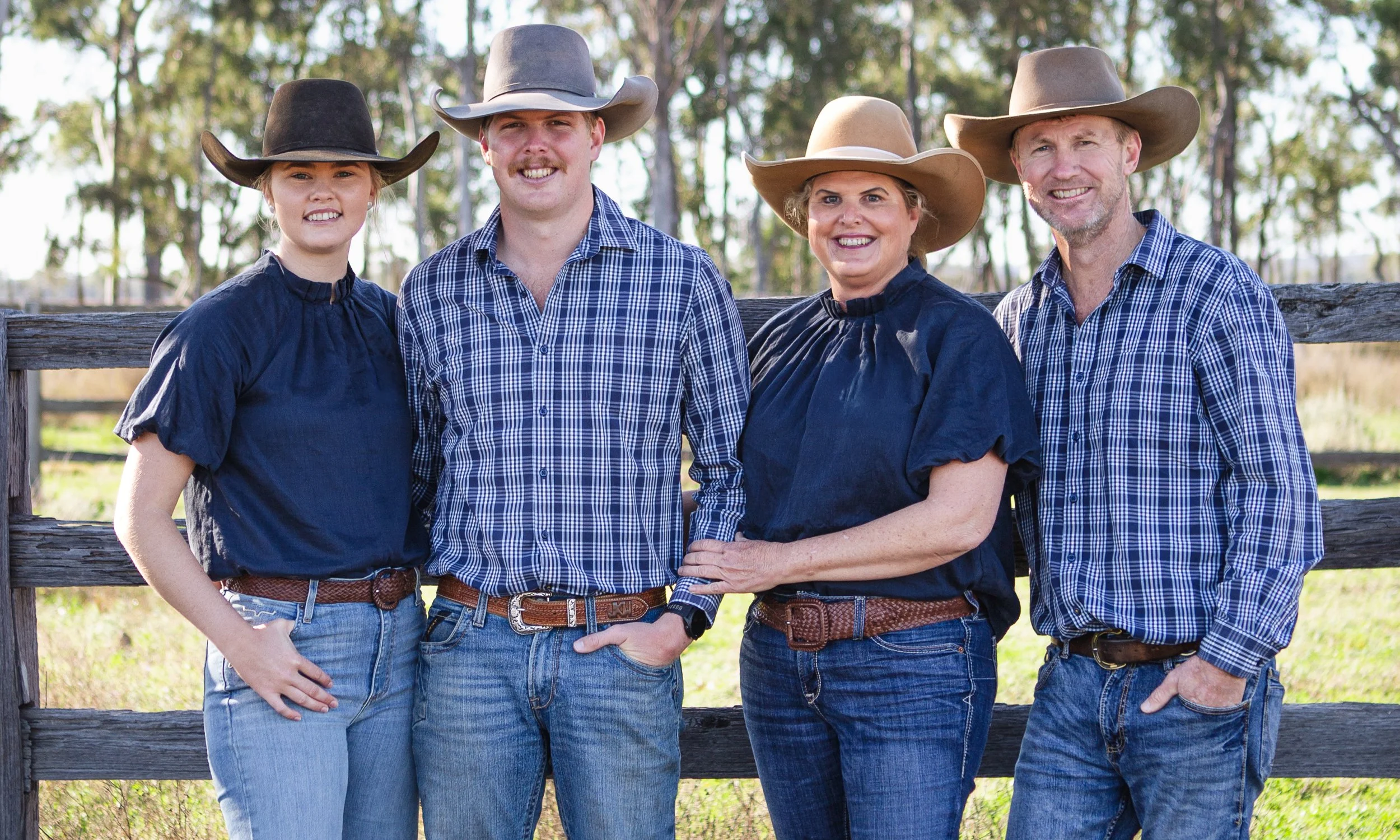 Family of four standing outdoors behind a wooden fence, dressed in plaid shirts and cowboy hats, smiling.