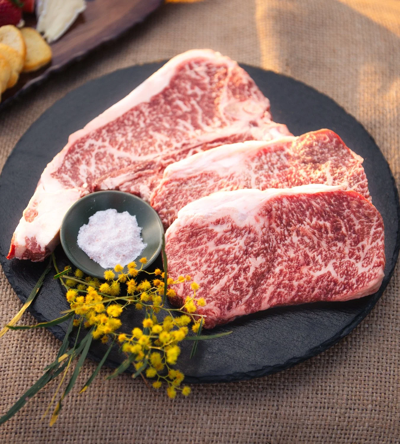 Three pieces of marbled raw beef steaks on a black slate serving platter with a small bowl of salt and yellow flowers for garnish.