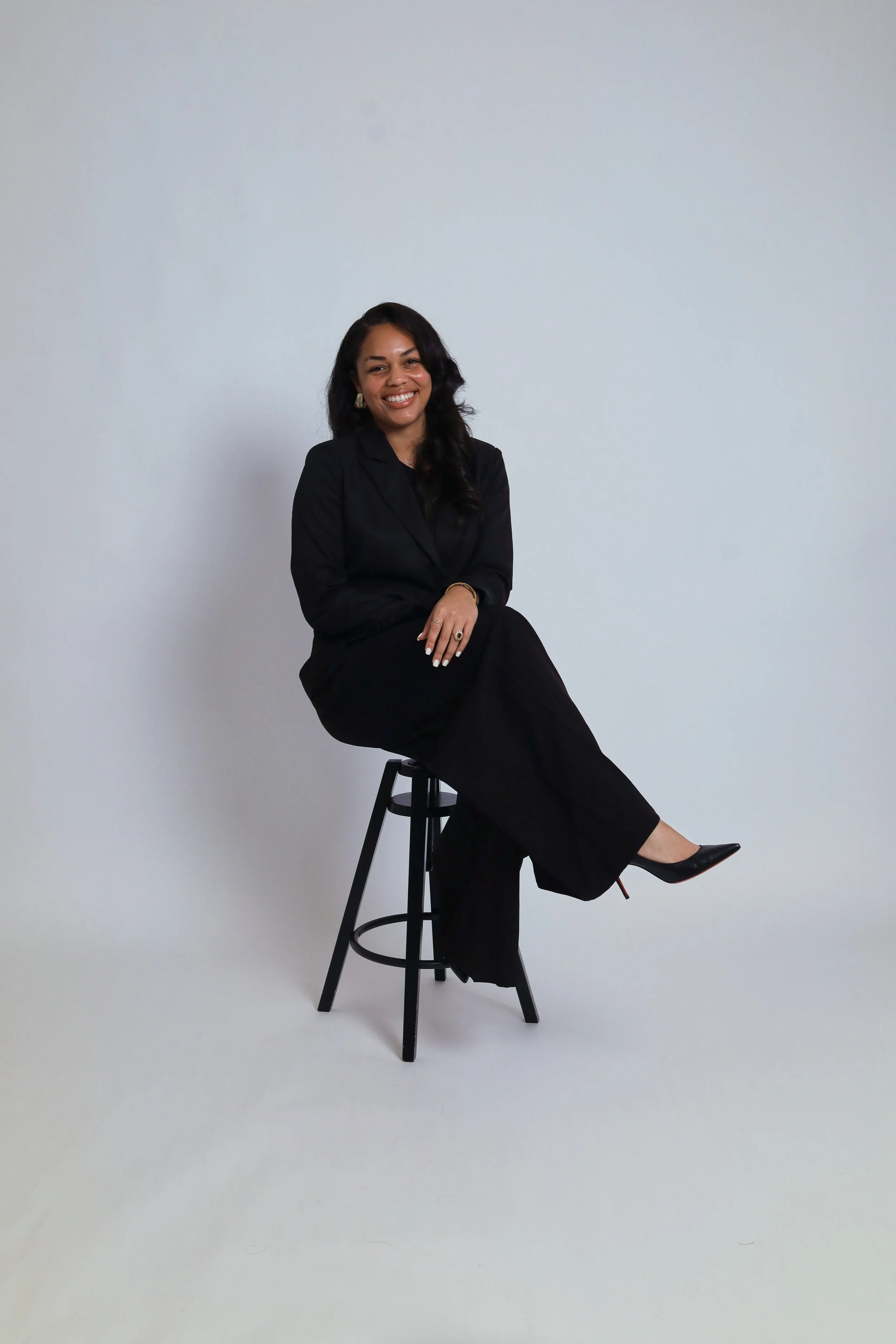 A woman with long dark hair and earrings, sitting on a black stool against a plain light gray background. She is smiling, dressed in a black suit and walking shoe, with her left arm resting on her lap and her right hand on her knee.