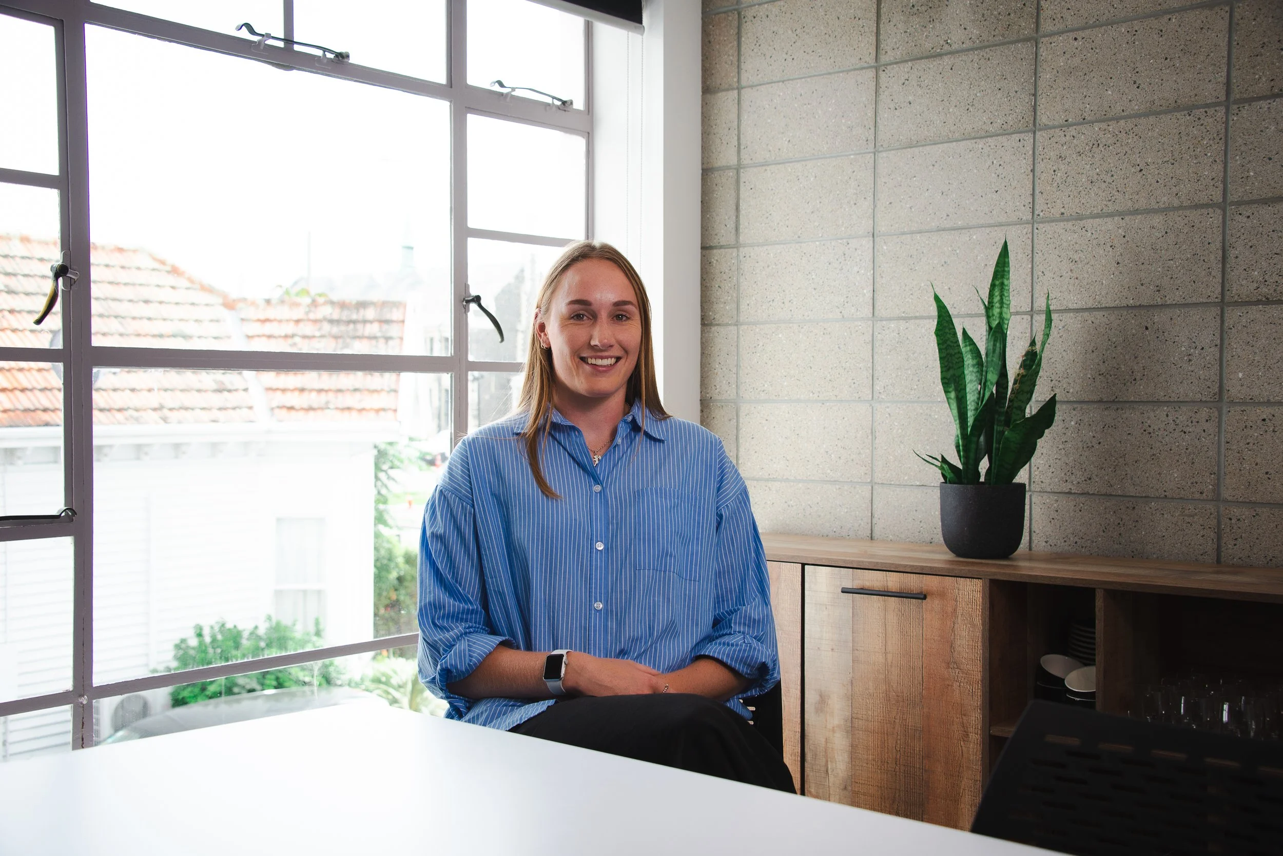 A woman with long blonde hair smiling while sitting at a table in a modern room with large windows, a potted plant, and a brick wall.