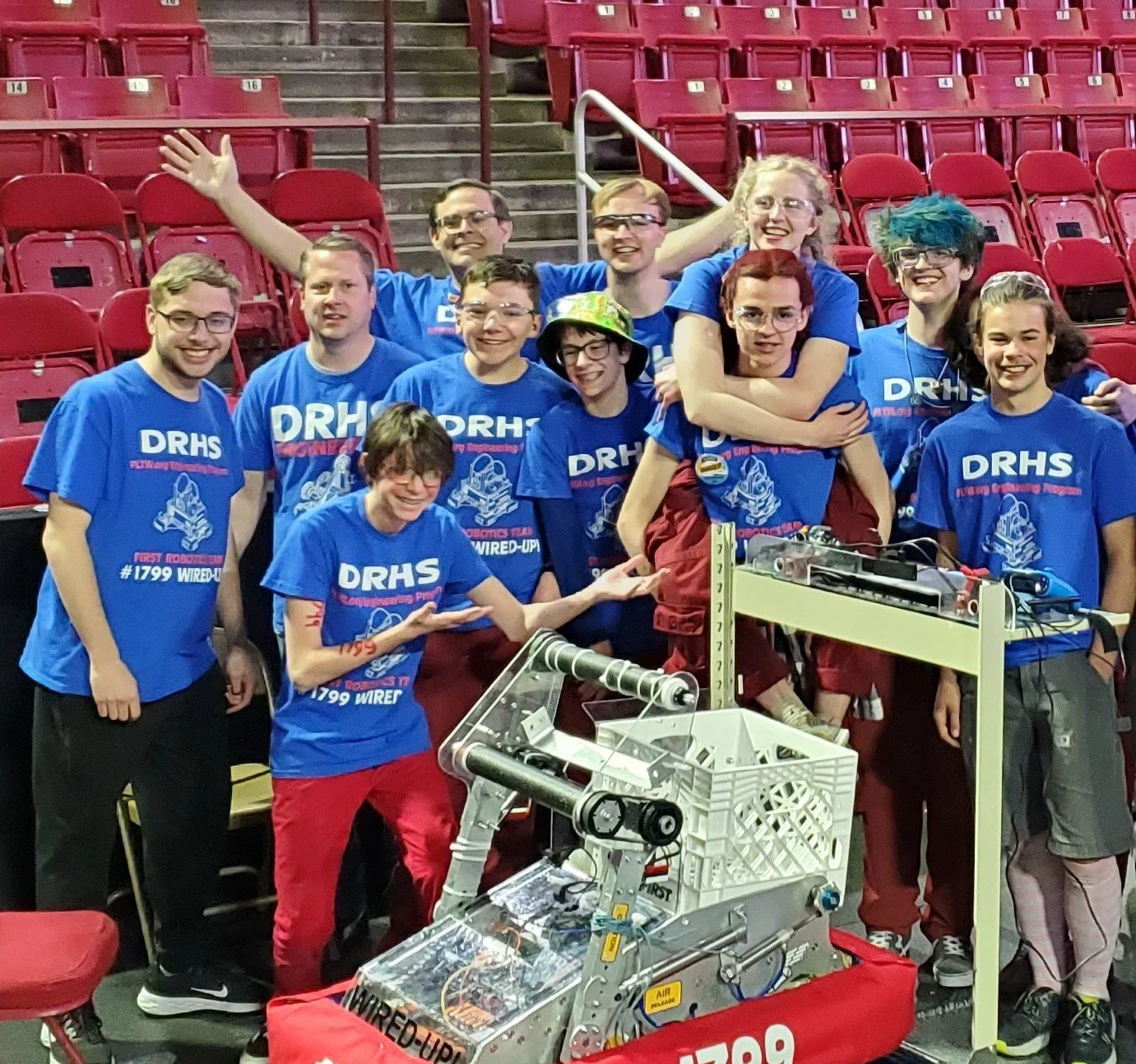 Group of robotics team members wearing blue T-shirts with 'DRHS' and robotics team logo, celebrating a 2023 Denver Regional win.