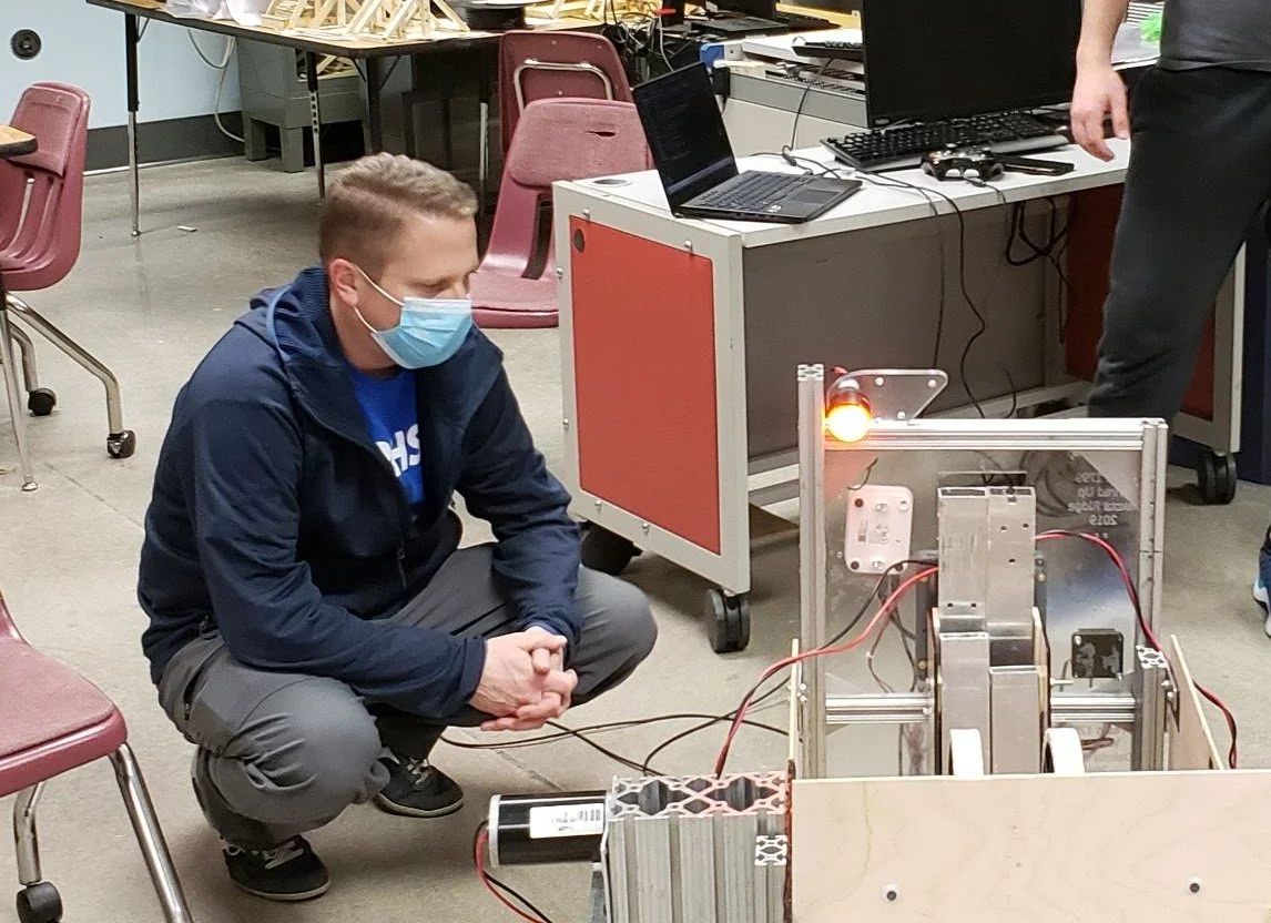Andy assessing the robot during the 2024 season. He's wearing a face mask crouching next to a robot with wires and a glowing red light in a classroom or lab setting.
