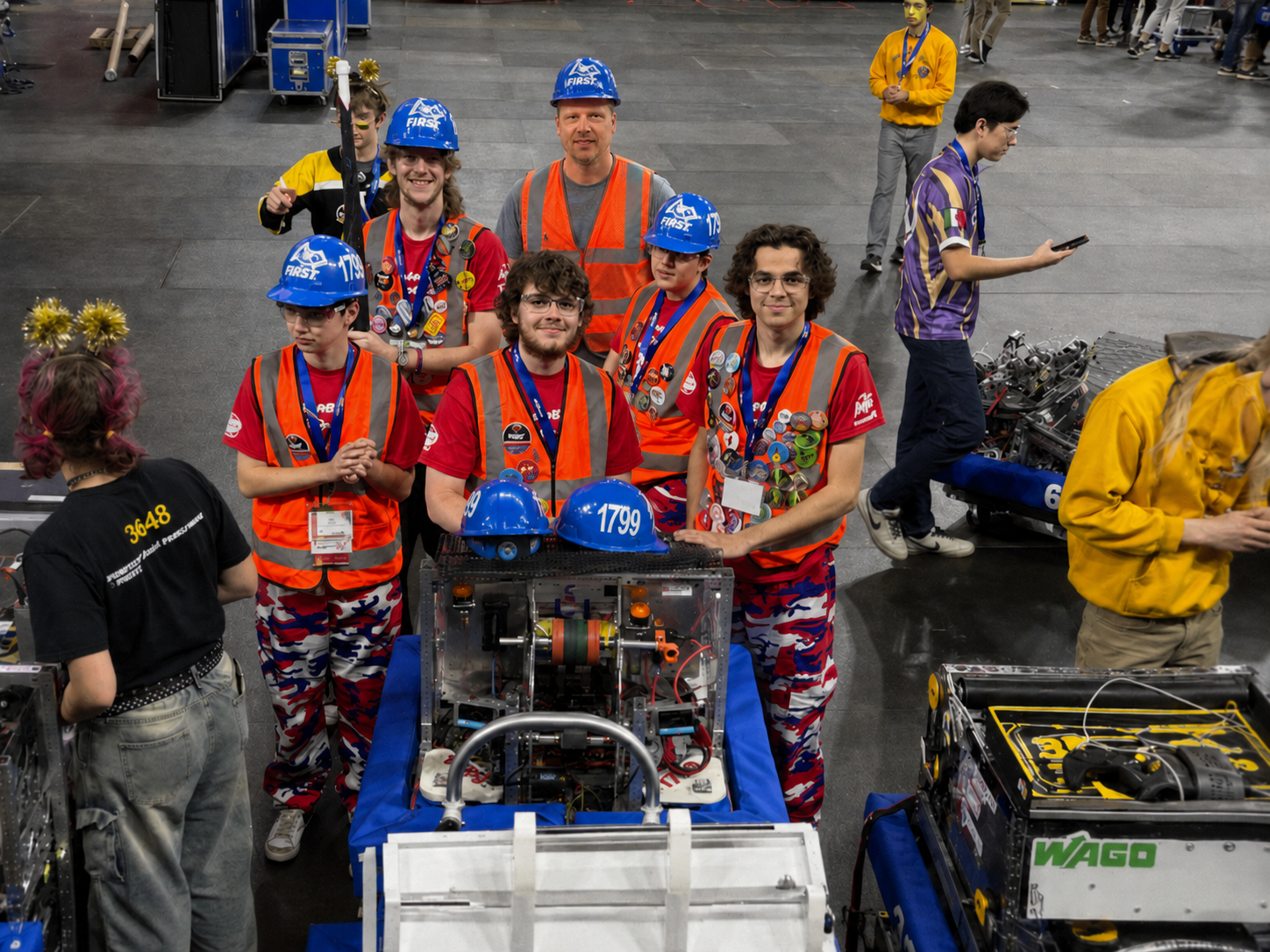 Group of robotics team members wearing orange safety vests and blue helmets with 'FIRST' logo, standing around their robot team at a competition, with medals around their necks.
